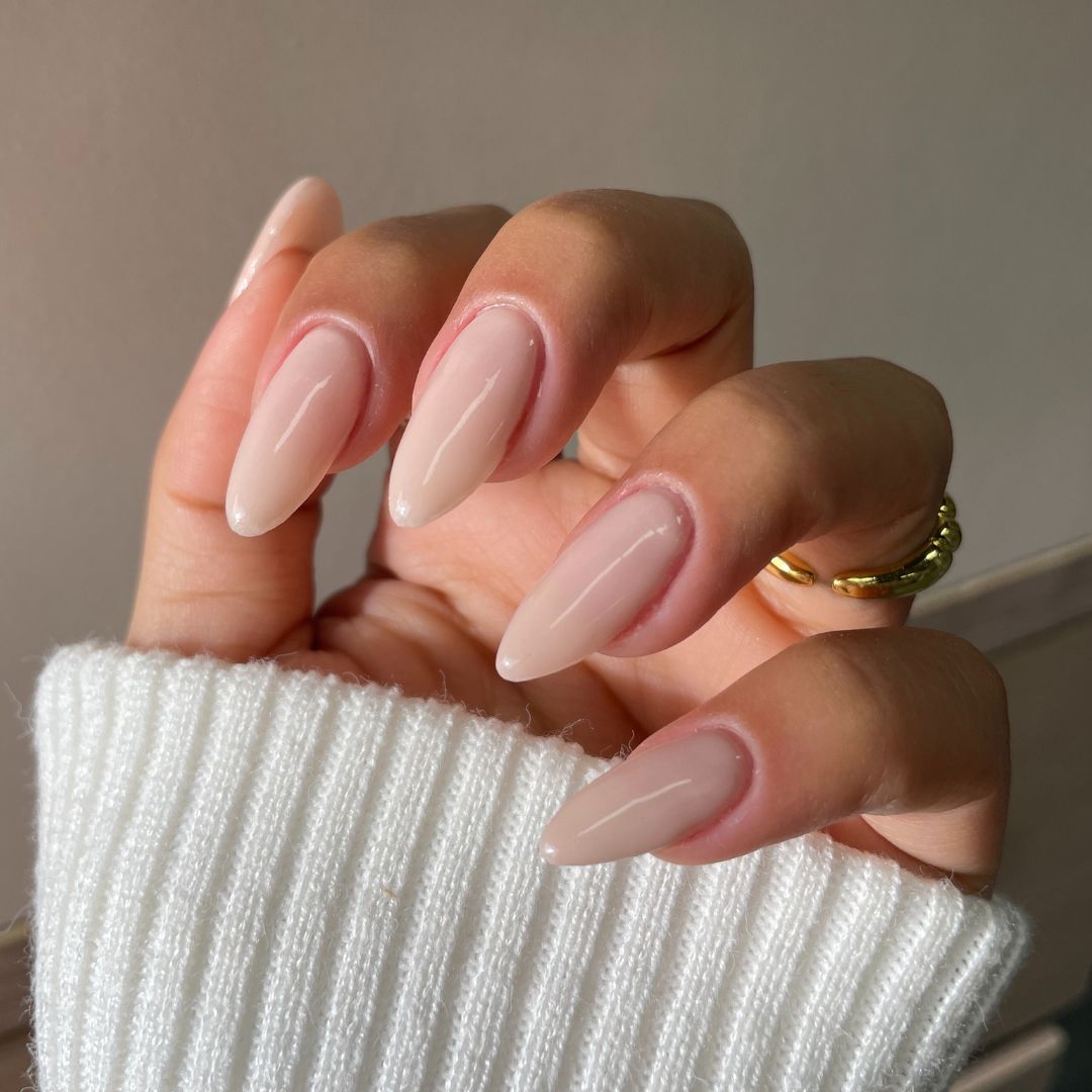 A hand displays almond-shaped nails coated in Sassy Saints' Sandstone Siren Dipping Powder, featuring glossy nude color, gold rings, and a white ribbed sweater sleeve partially covering the fingers.