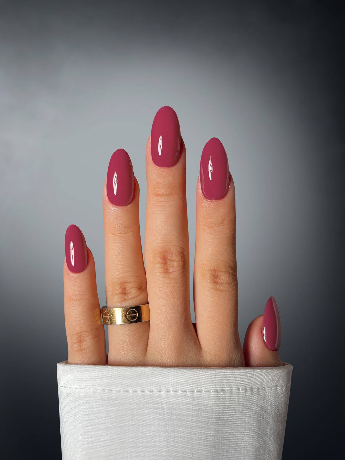 A hand with long, oval glossy pink nails created using Sassy Saints Cherry Charmour Dipping Powder, wearing a gold ring and partially covered by a white shirt sleeve, is set against a gradient gray background.