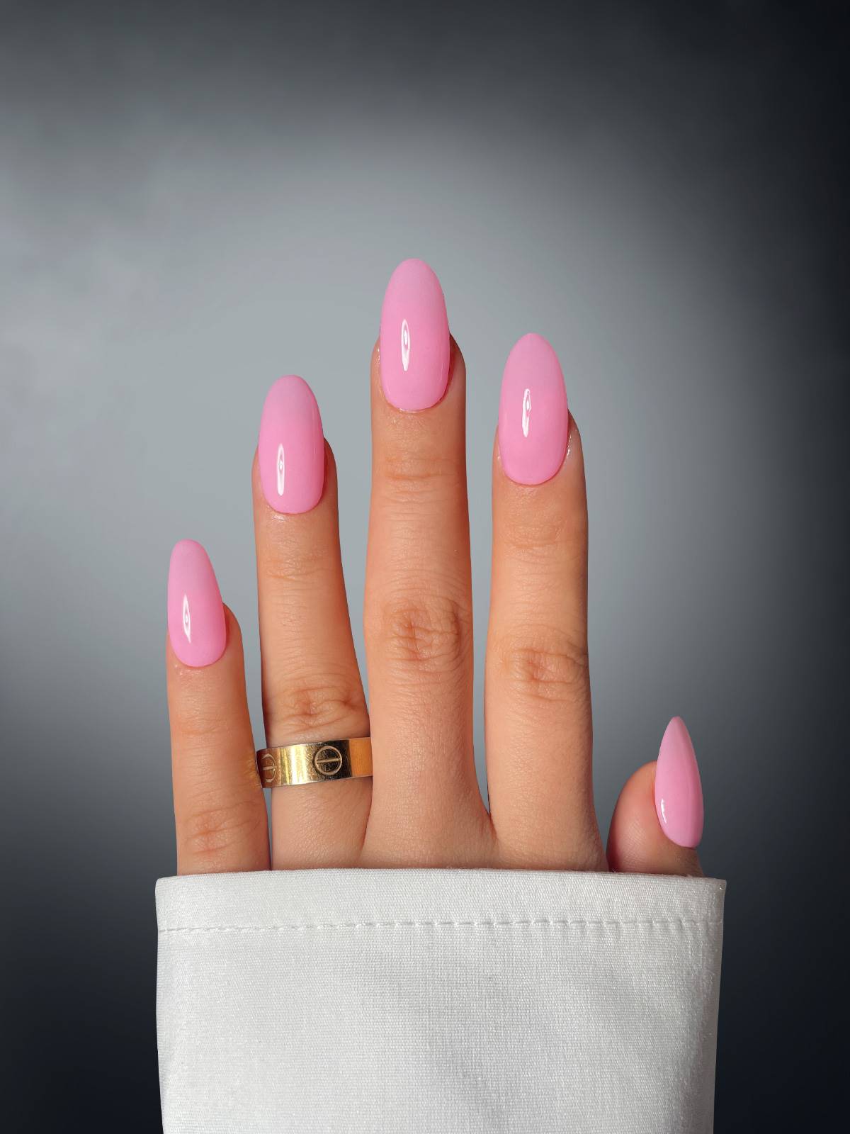 Four-fingered hand with long, glossy pink nails coated in Sassy Saints' Cotton Candy Cutie Dipping Powder. A gold ring adorns the middle finger, partially covered by a white sleeve against a dark gradient background.