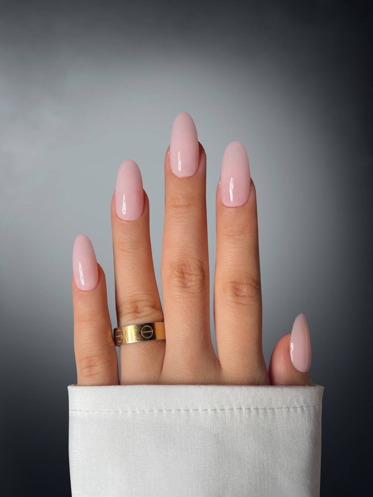 A hand with glossy light pink nails, colored with Sassy Saints - Brings The Salon Home's Pearly Pursuader Dipping Powder, wears a gold ring on the middle finger. A white shirt sleeve partially covers the fingers against a smooth gradient background.