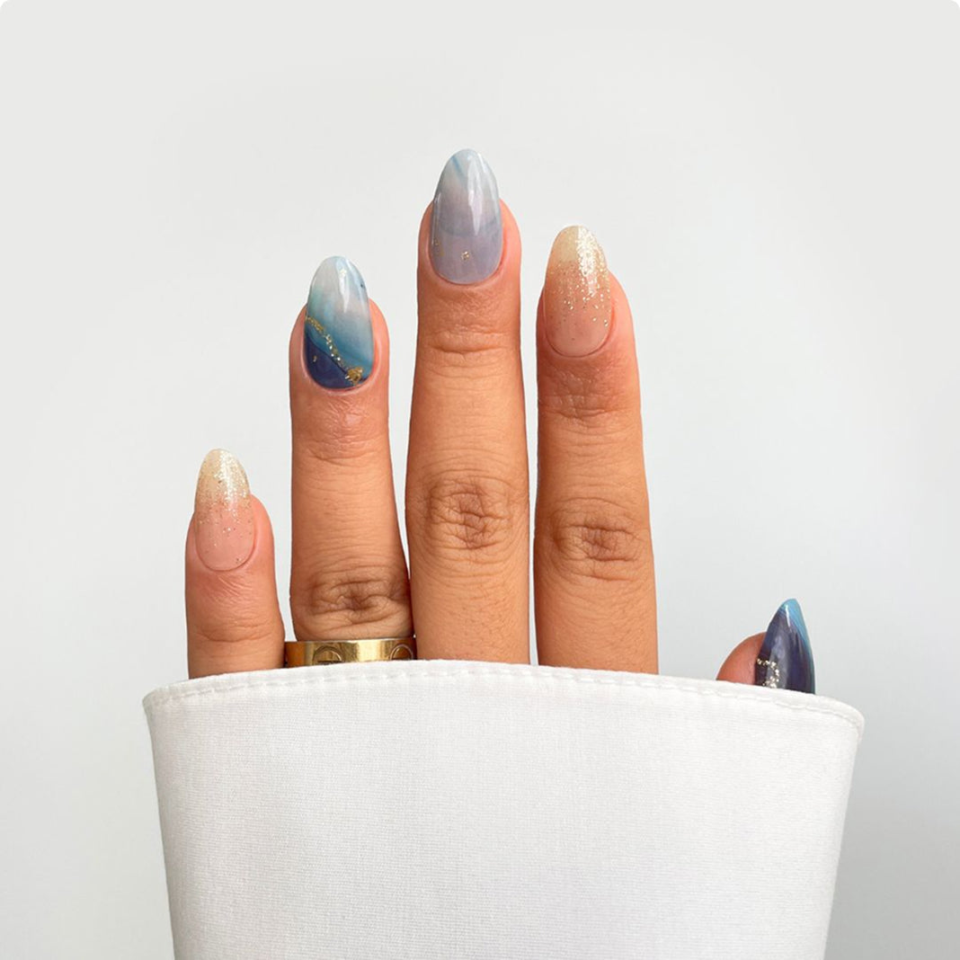 A hand with long, almond-shaped nails showcases Sassy Saints Ocean Dream Gel Wrap—blue, gold, and nude marble designs with glitter accents—paired with a gold ring and white fabric against a plain background.