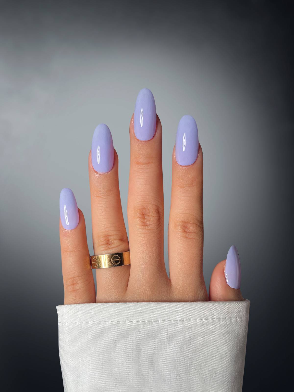 A hand with five fingers features Sassy Saints - Brings The Salon Home’s Lavender Luxe Dipping Powder and a gold ring on the middle finger, set against a gradient gray background. A white shirt sleeve partially covers the wrist.