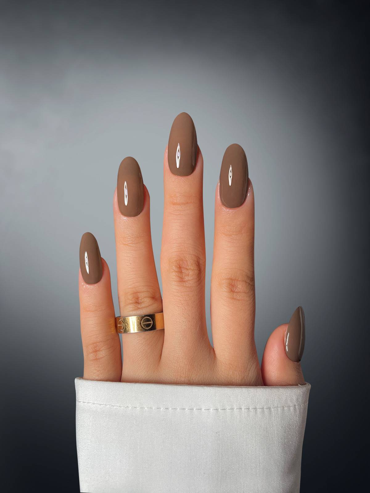 A hand showcases Cocoa Cutie Dipping Powder by Sassy Saints - Brings The Salon Home, with brown oval nails adorned with small white eye designs. The person wears a gold ring and white long sleeve; the background is a smooth dark-to-light gradient.