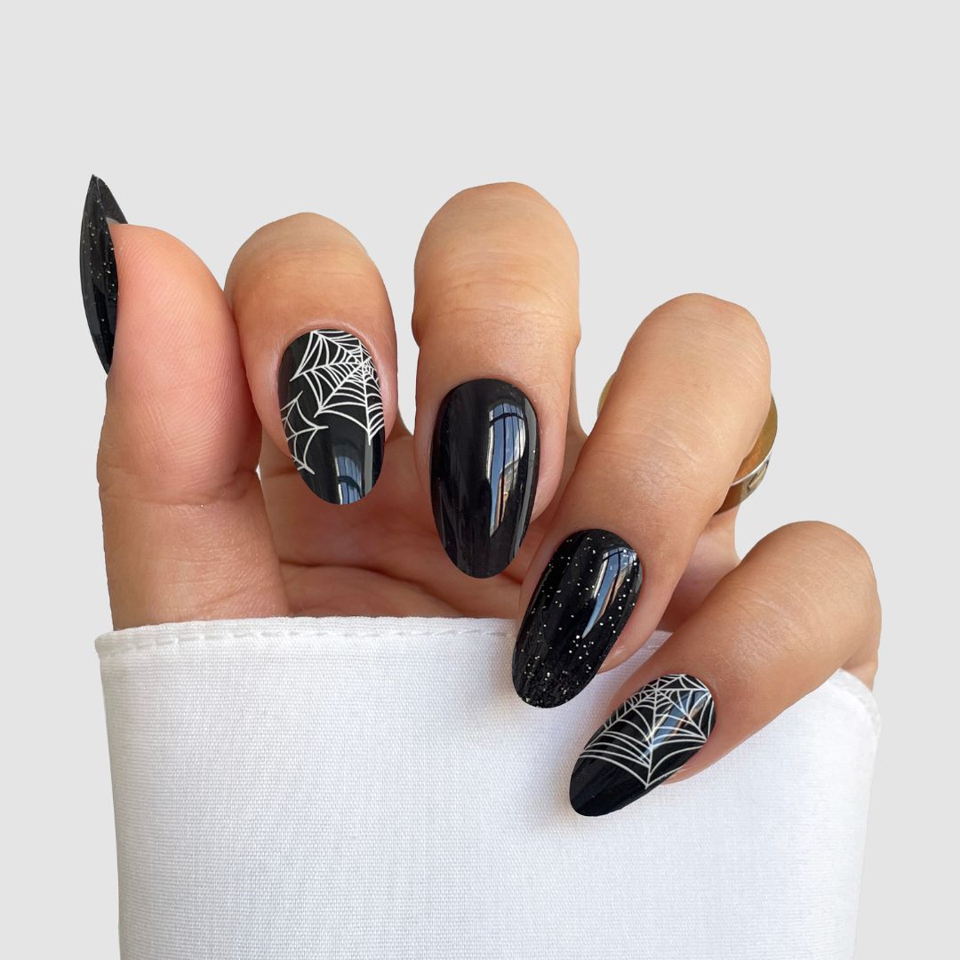 A hand wears Sassy Saints’ Webbed Luxe Gel Wrap: black, almond-shaped nails with white spider webs on two and white speckles on the rest. The look is set against a light gray background, with a white sleeve partially covering the hand.