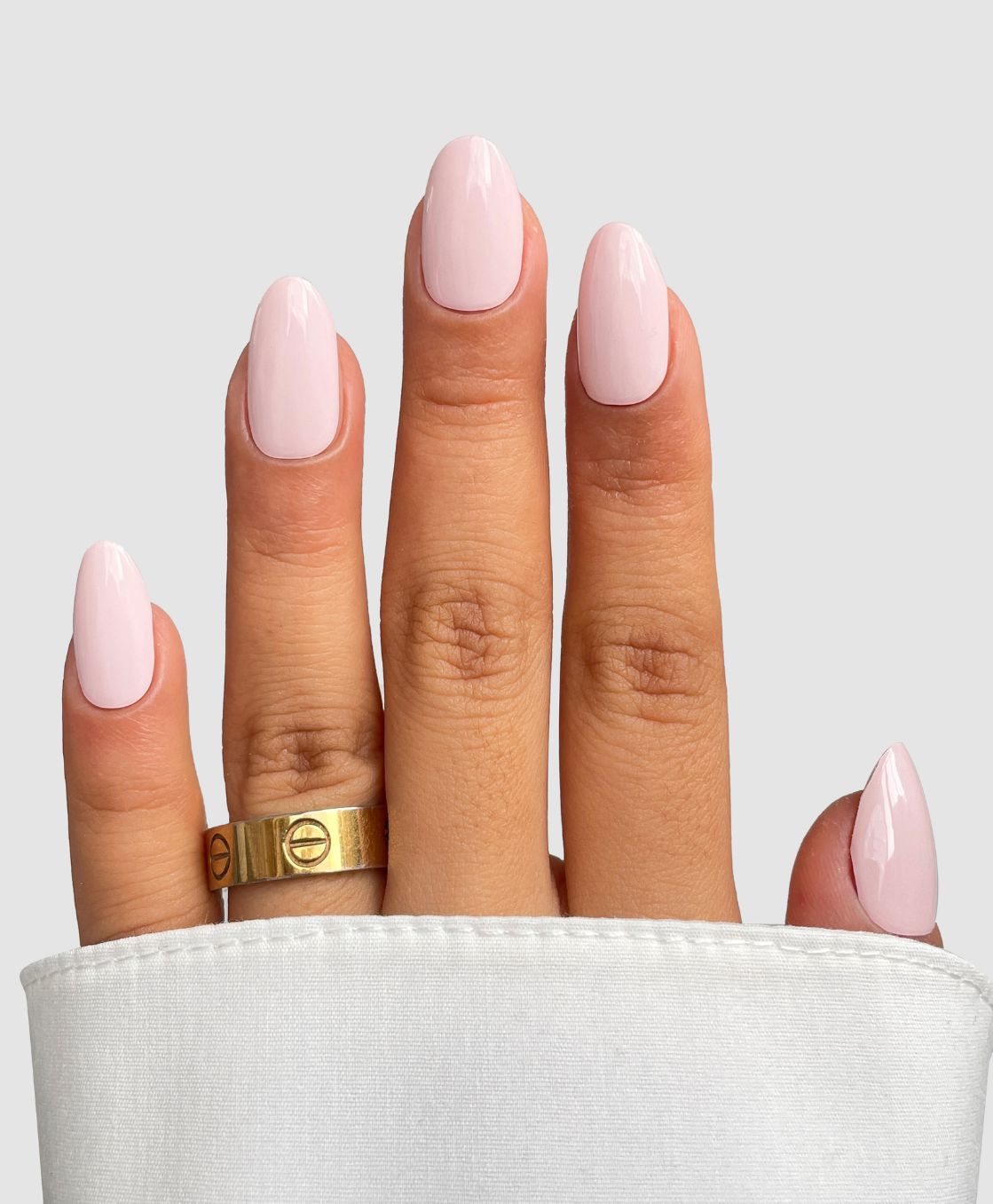 A hand showcases Sassy Saints' Marshmallow Drip Gel Wrap gel nails in light pink almond shapes, with two gold rings on the middle finger and a white sleeve at the wrist, set against a light gray background.