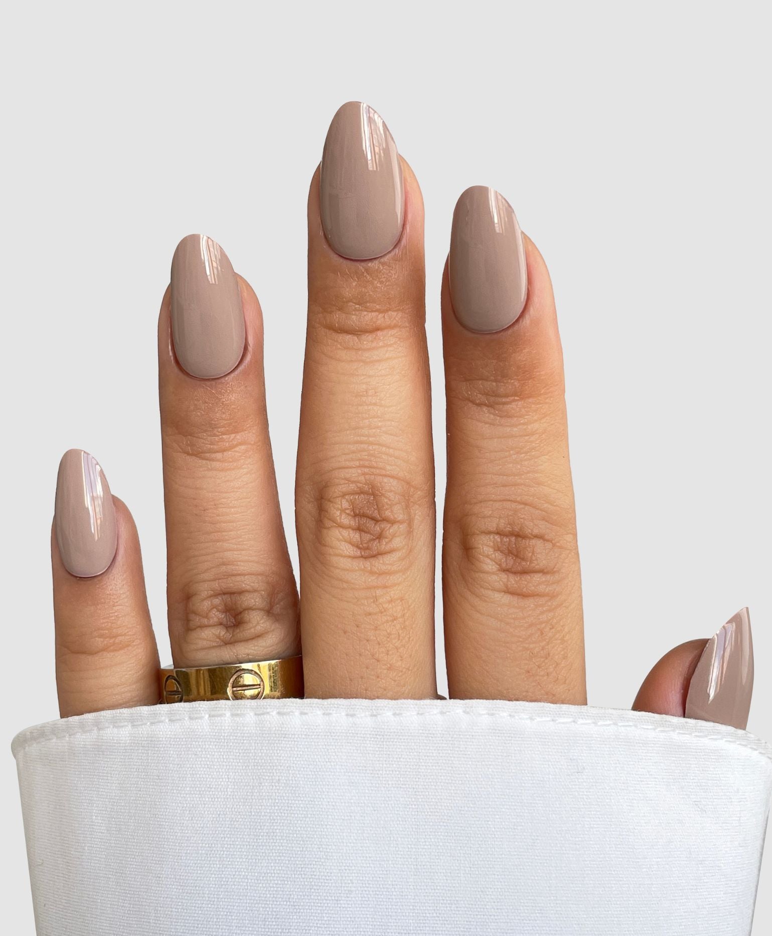A neatly manicured hand wears Sassy Saints - Brings The Salon Home’s Dusty Stone Gel Wrap, a glossy taupe shade. A gold ring adorns the finger, with the hand part-wrapped in white fabric against a plain light backdrop.