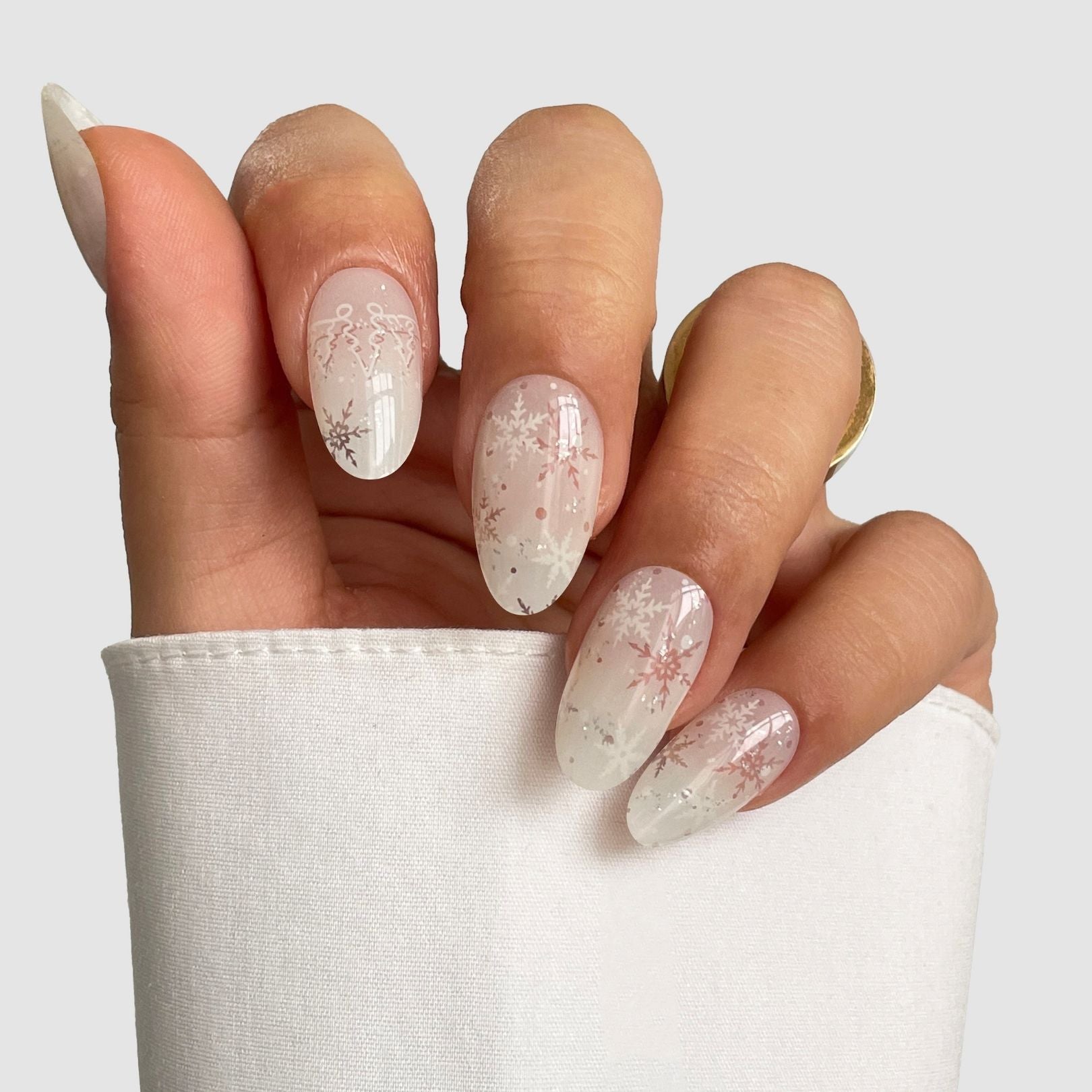 A hand with long, almond-shaped Arctic Frost Gel Wrap nails by Sassy Saints—featuring a translucent nude base and white snowflake designs—is shown against a light gray background. The wearer has a white cuffed sleeve and gold ring.