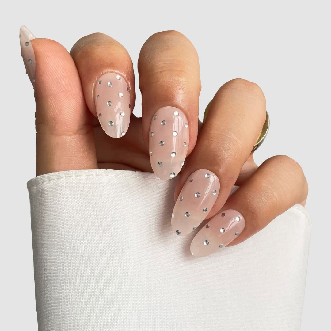 A hand with long, oval nails features Sassy Saints - Brings The Salon Home’s Platinum Spotlight Gel Wrap, a sheer nude finish with silver rhinestone accents, displayed against a white fabric background.