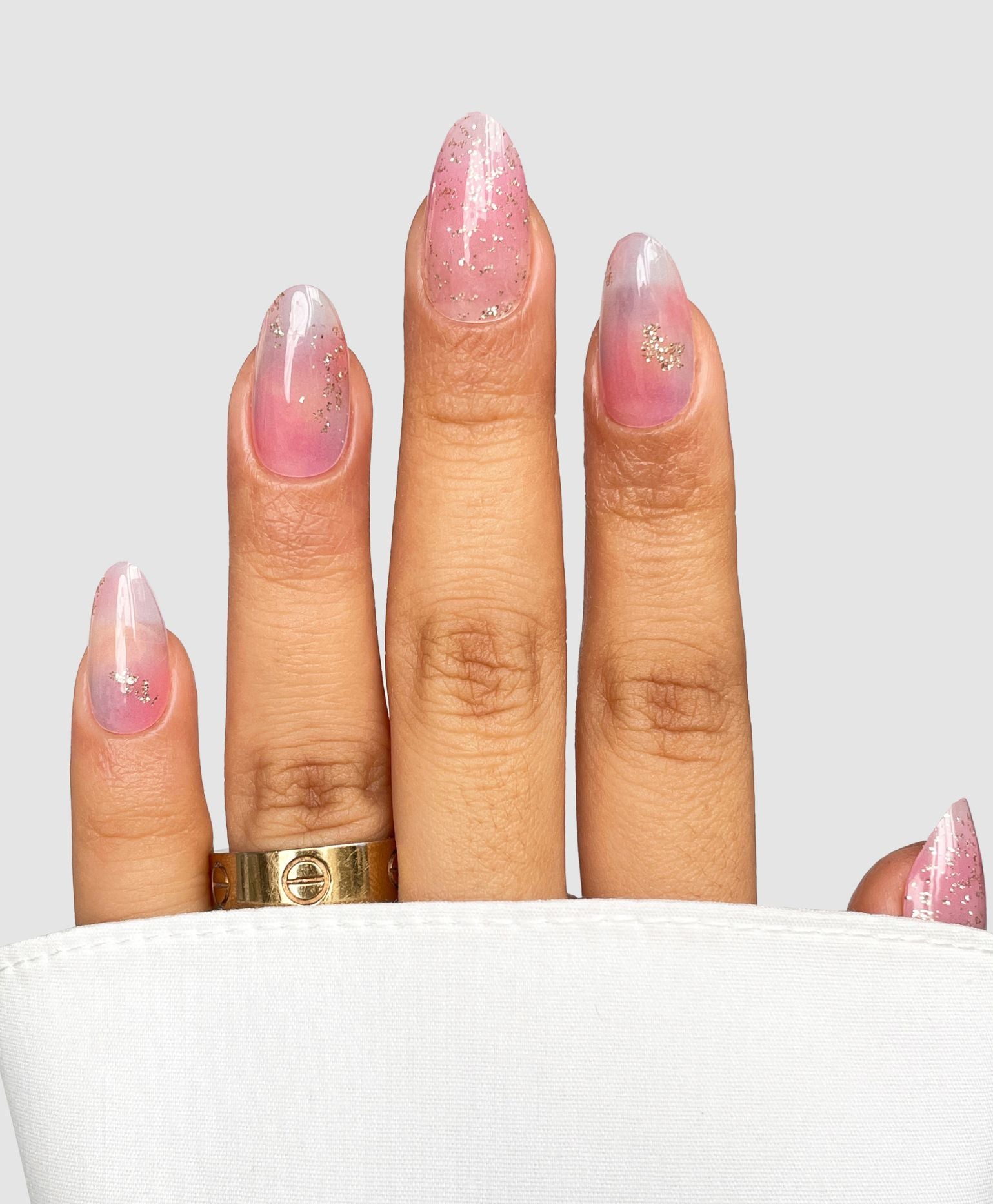 A hand with long, almond-shaped nails features Sassy Saints' Pastel Haze Gel Wrap in a pink ombre with glitter accents. The look is complemented by a gold ring and displayed on a white cloth against a light gray background.