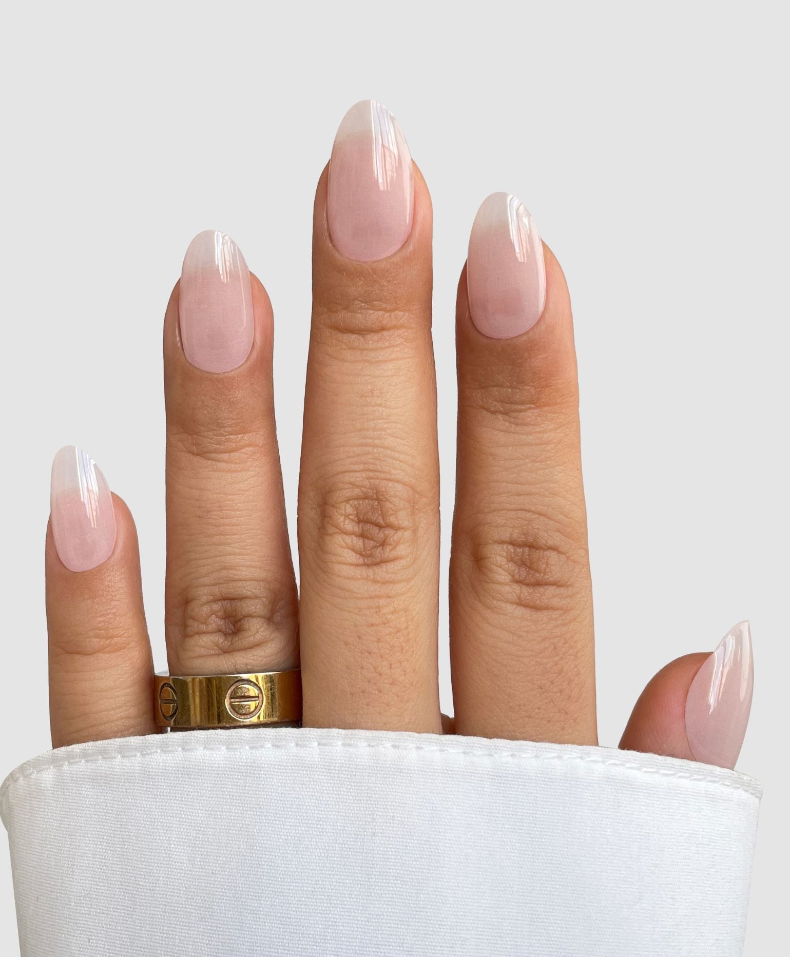 A hand with medium-length, almond-shaped nails showcases Sassy Saints’ Sugar Glaze Gel Wraps—a light pink manicure with white tips. A gold ring adorns the middle finger and a white cuff partially covers the hand.