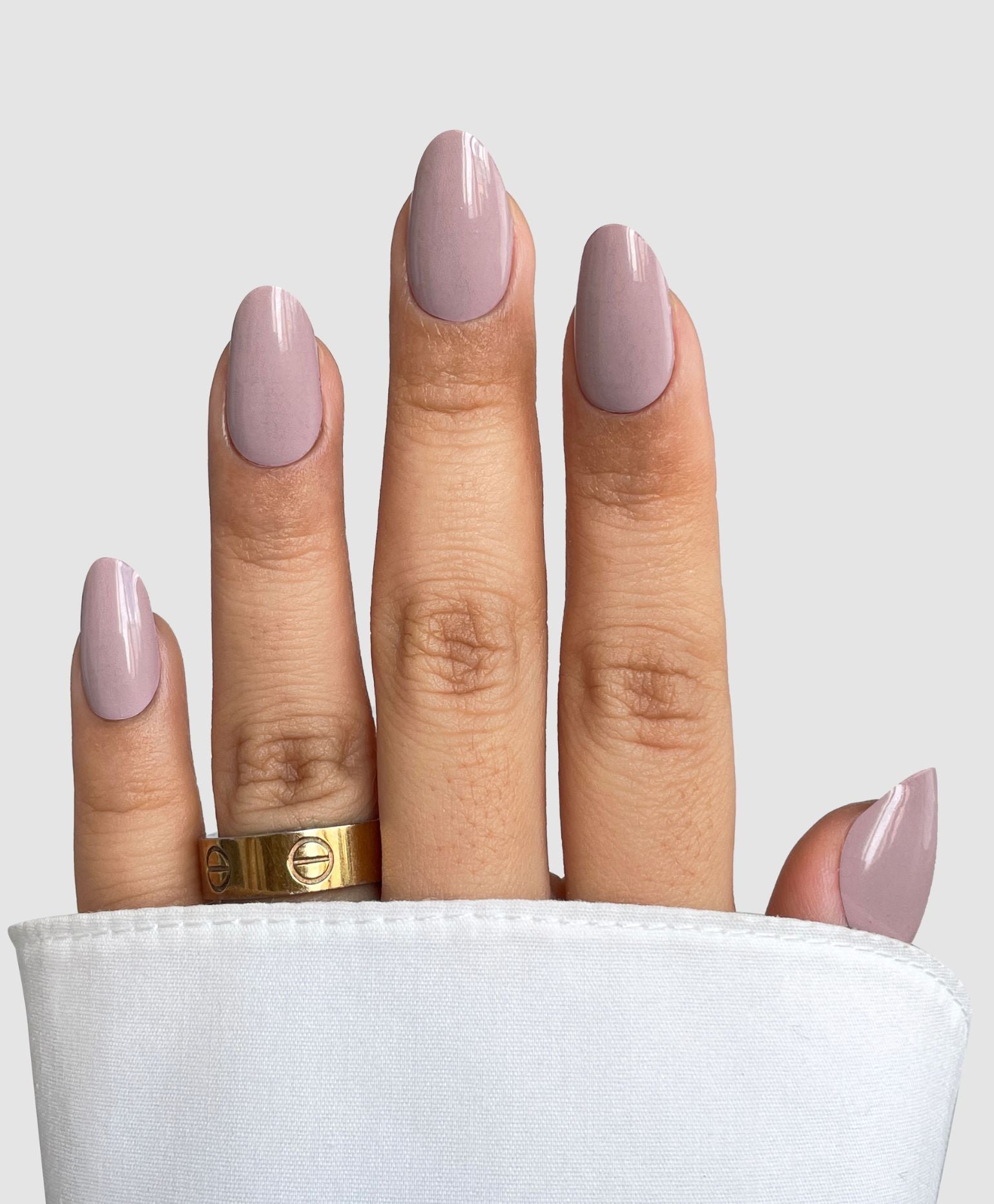 A hand with neatly manicured, almond-shaped nails features Sassy Saints - Brings The Salon Home’s Neutral Flow Gel Wrap in soft mauve. Two gold rings adorn the middle finger and a white sleeve partially covers the hand against a light gray background.
