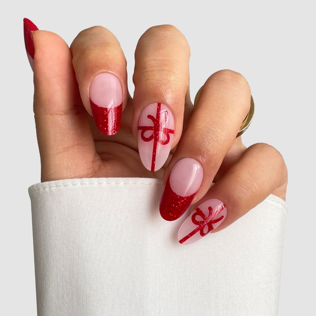 A hand with long oval nails sports Sassy Saints' Gift Wrapped Gel Wrap, featuring a red and pink manicure with red glitter tips, lines, and bow designs like wrapped gifts. A white sleeve partly covers the hand.
