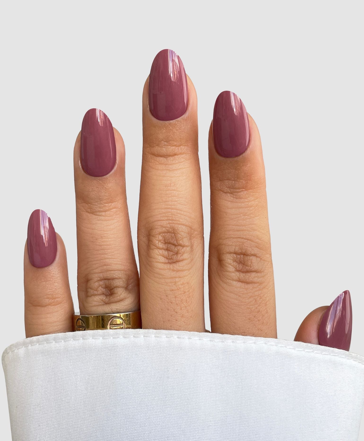A hand features long, oval-shaped nails adorned with Sassy Saints' Deep Petal Gel Wrap. The mauve finish pops next to a gold ring, white cuffed sleeve, and a neutral gray background—Brings The Salon Home.
