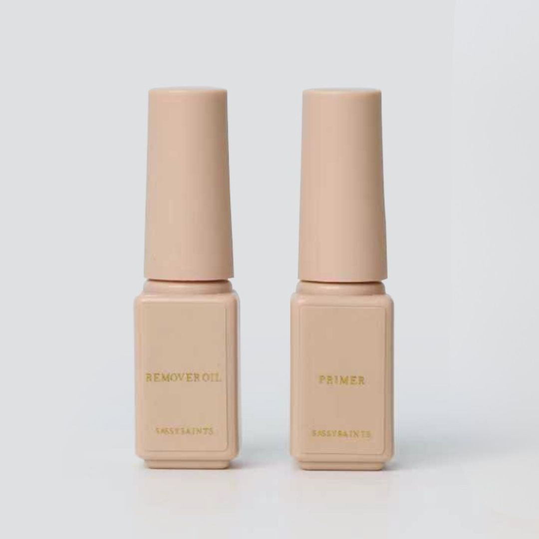 Two beige bottles labeled "Remover Oil" and "Primer" in gold text, from Sassy Saints' Primer & Remover Oil Bundle (FREE), stand side by side against a plain light gray background.