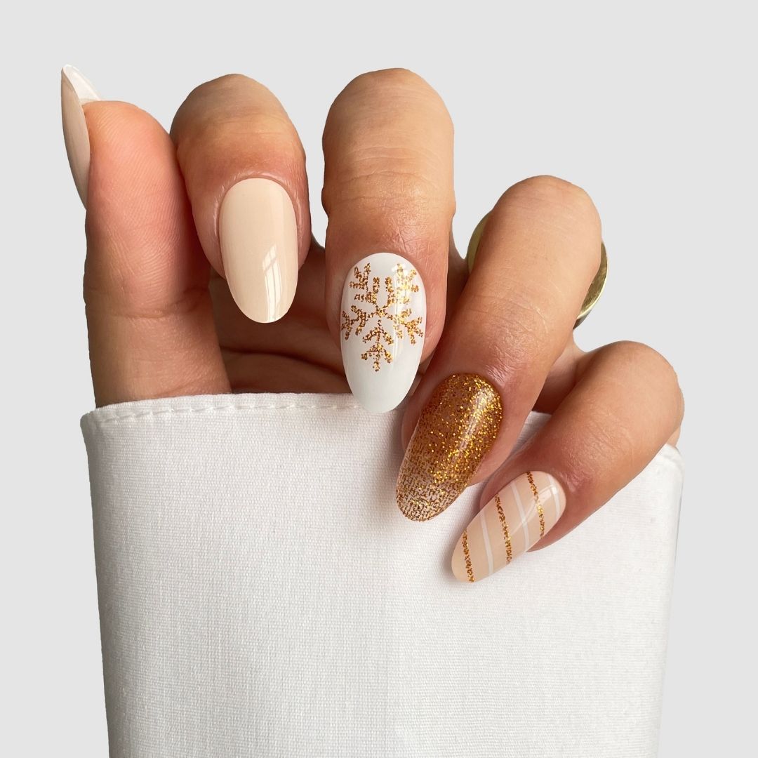 A hand showcases Sassy Saints' Golden Flurry Gel Wraps—almond-shaped nails in beige, white, and gold with designs like a gold snowflake, glittery gold accents, and stripes—partially wrapped in white fabric.