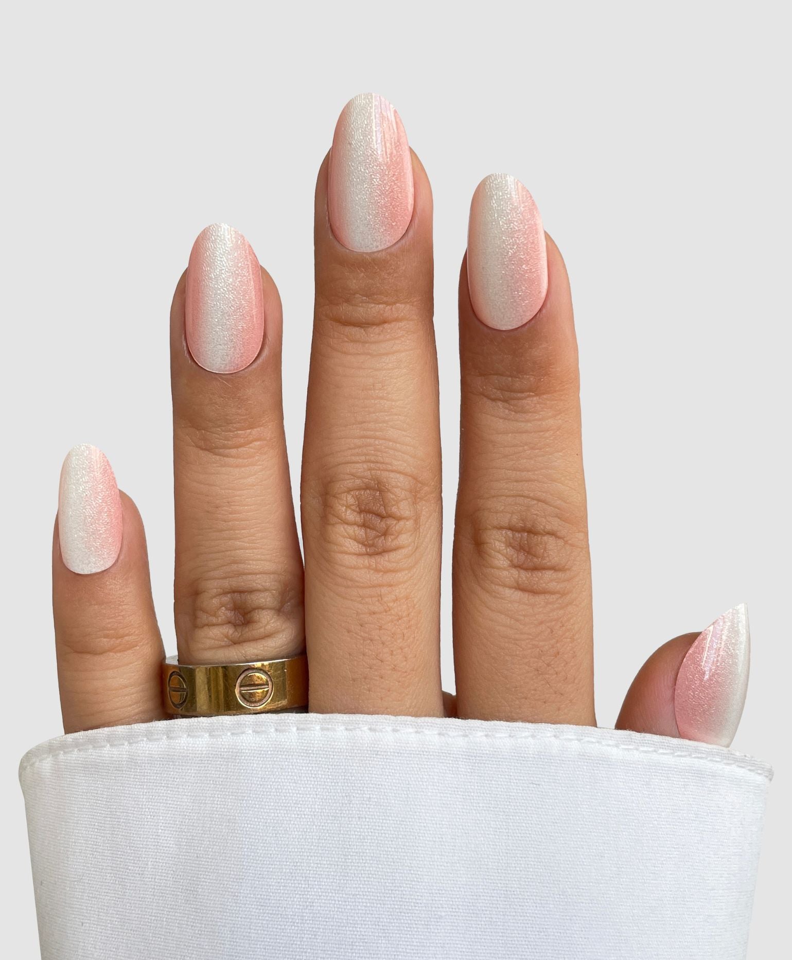 A hand with medium-length oval nails shows off Sassy Saints - Brings The Salon Home's Silk Finish Gel Wrap in a white ombré shimmer. Two gold rings adorn the ring finger, a white cuff covers part of the hand, and the background is light gray.