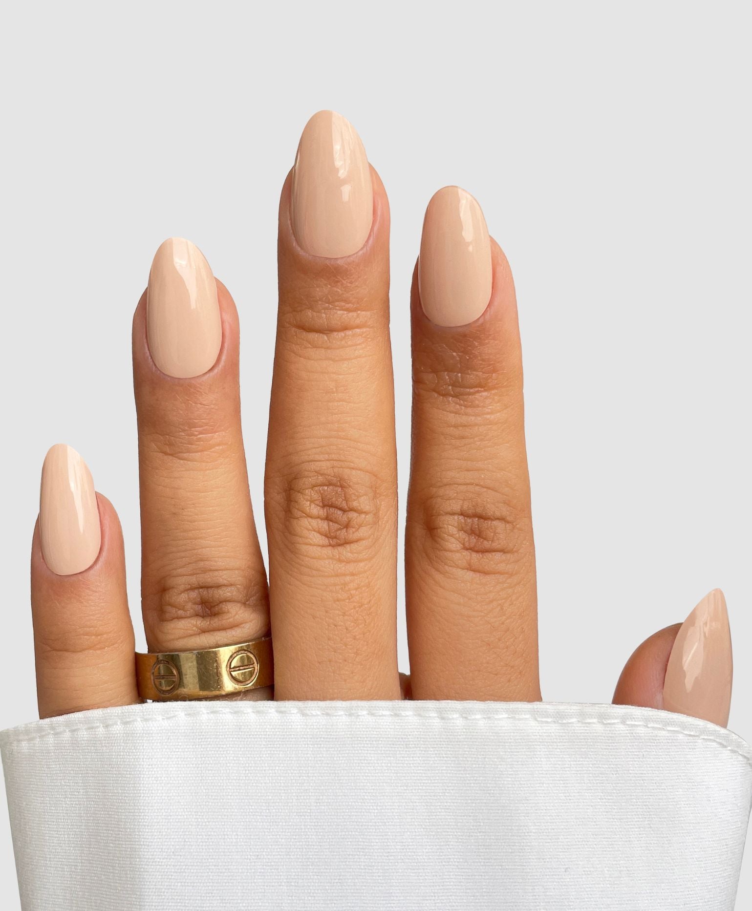 A hand with almond-shaped nails coated in Sassy Saints - Brings The Salon Home's Naked Ambition Gel Wrap, accented by a gold ring on the middle finger and partially draped in white fabric.