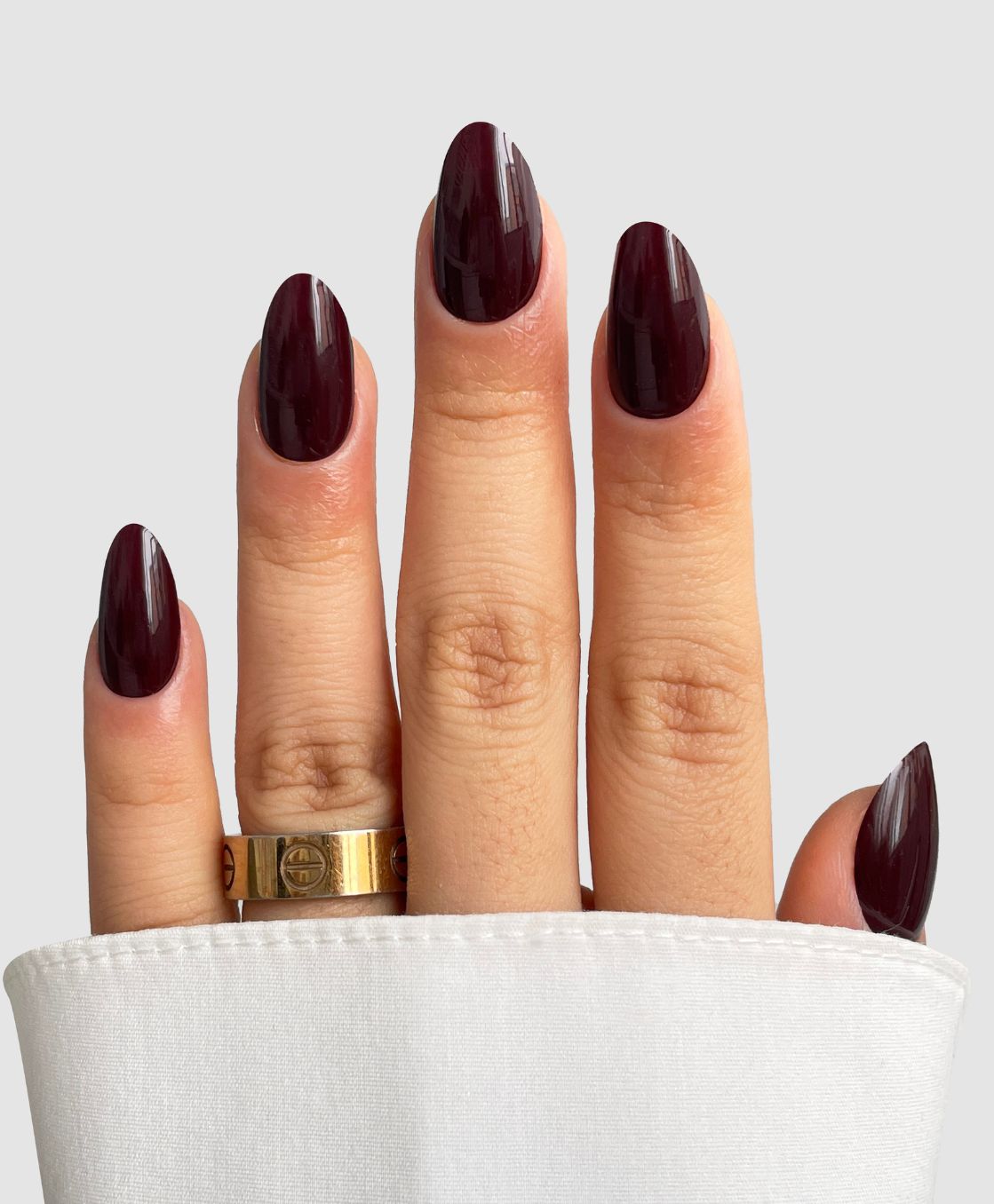 A hand with Sassy Saints - Brings The Salon Home's Dark Current (Dark Red) almond-shaped nails is shown against a light background, adorned with a gold ring and a white cuffed sleeve.