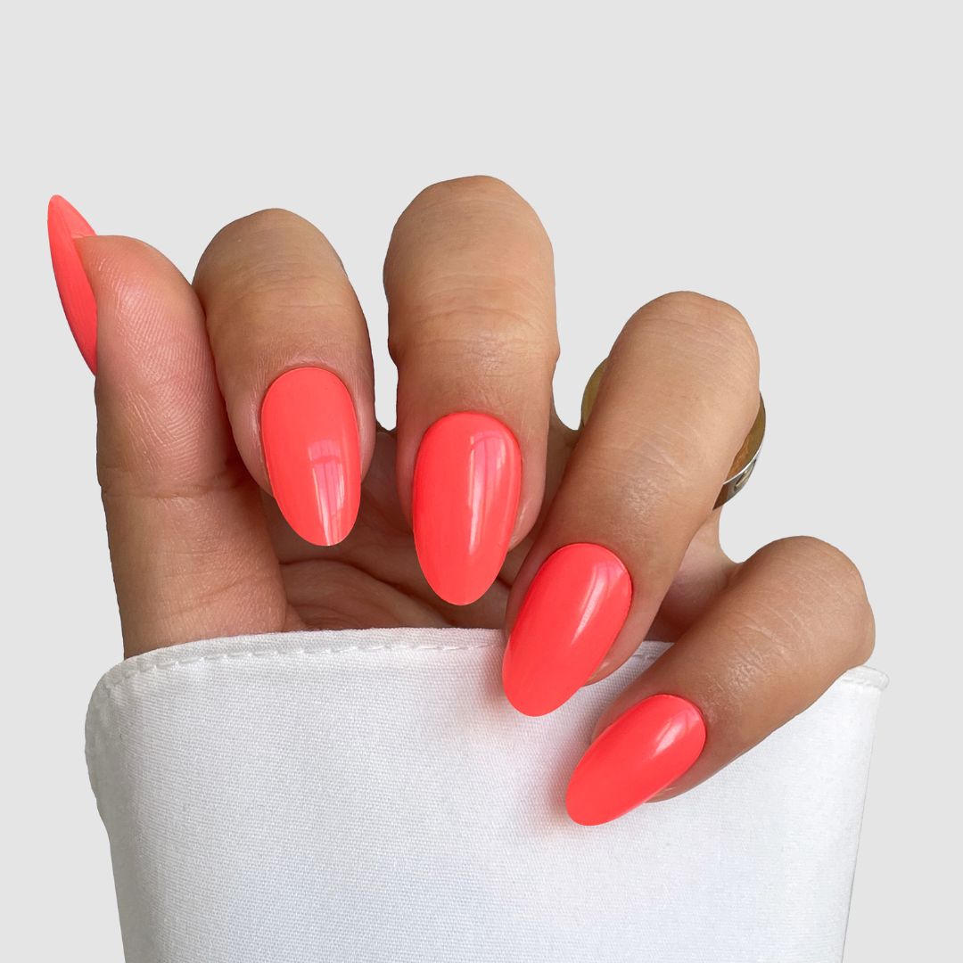A hand with long, almond-shaped nails in Sassy Saints' Coral Kiss Gel Wrap, a glossy neon coral shade, partially covered by a white sleeve against a light gray background.