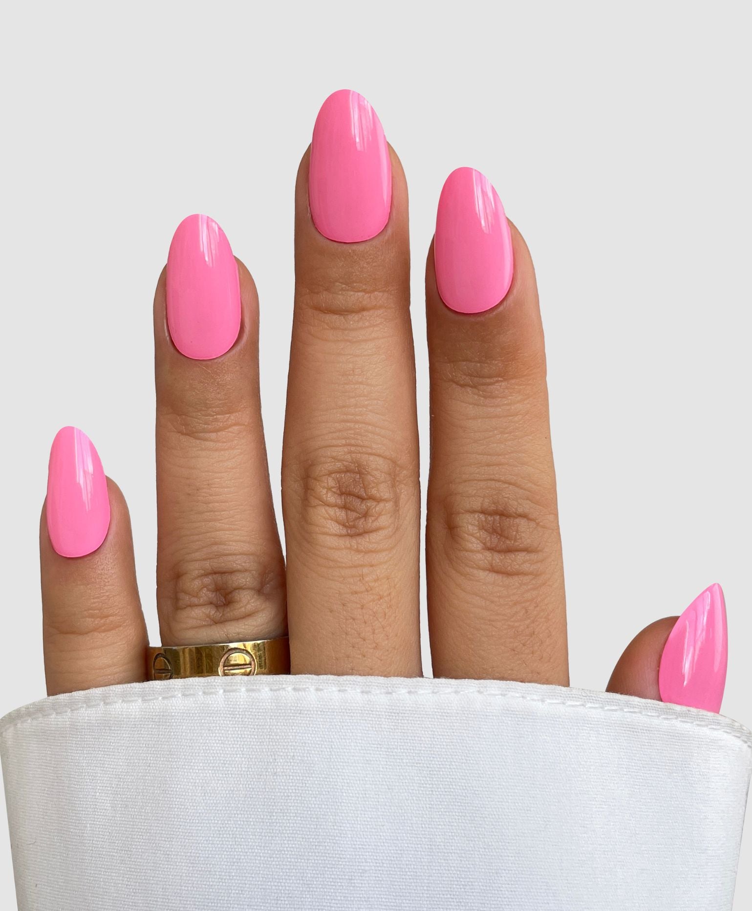 A hand with neatly manicured nails in bright pink Sweet Beat Gel Wrap by Sassy Saints - Brings The Salon Home, partially covered by a white sleeve. A gold ring adorns the ring finger, set against a plain, light-colored background.