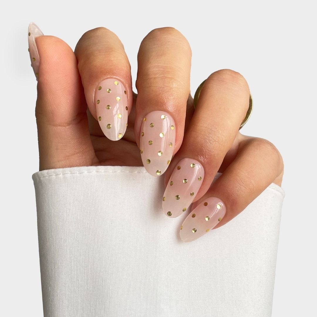 A hand with long, almond-shaped nails wearing Sassy Saints - Brings The Salon Home's Champagne Drops Gel Wrap, featuring sheer nude with gold dots. The person wears a white long-sleeve top and a gold ring.