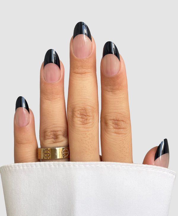 A hand with almond-shaped nails wears Sassy Saints' Midnight French Gel Wraps—a black French tip manicure—plus two gold rings, partially covered by a white sleeve on a light gray background.