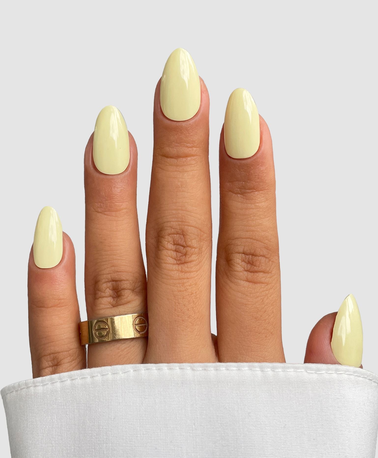 A hand with long, almond-shaped nails wears Buttercup Yellow Gel Wrap by Sassy Saints - Brings The Salon Home. A gold ring adorns the middle finger, a white sleeve partly covers the hand, and a light gray background completes the look.