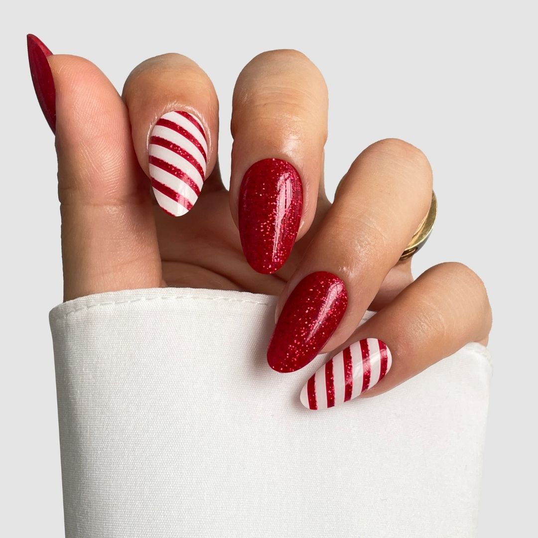 A hand with almond-shaped nails features Sassy Saints' Candy Cane Gel Wraps: three nails in sparkly red and two with red-and-white candy-cane stripes, all partially covered by a white sleeve. Brings the salon home for festive style.