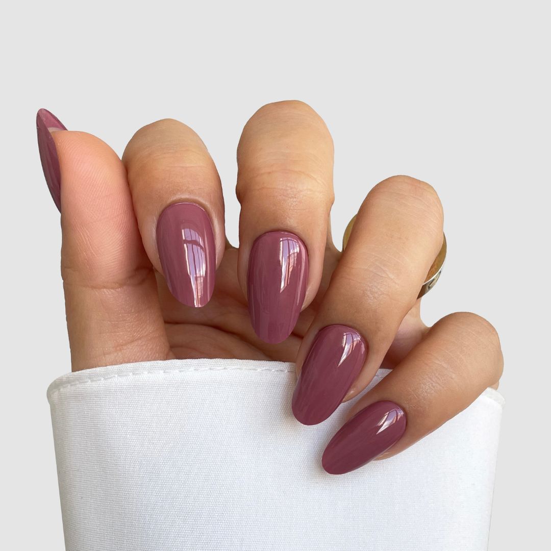 A hand with long, almond-shaped nails features Sassy Saints’ Deep Petal Gel Wrap in glossy mauve, resting against a white sleeve on a light gray background.