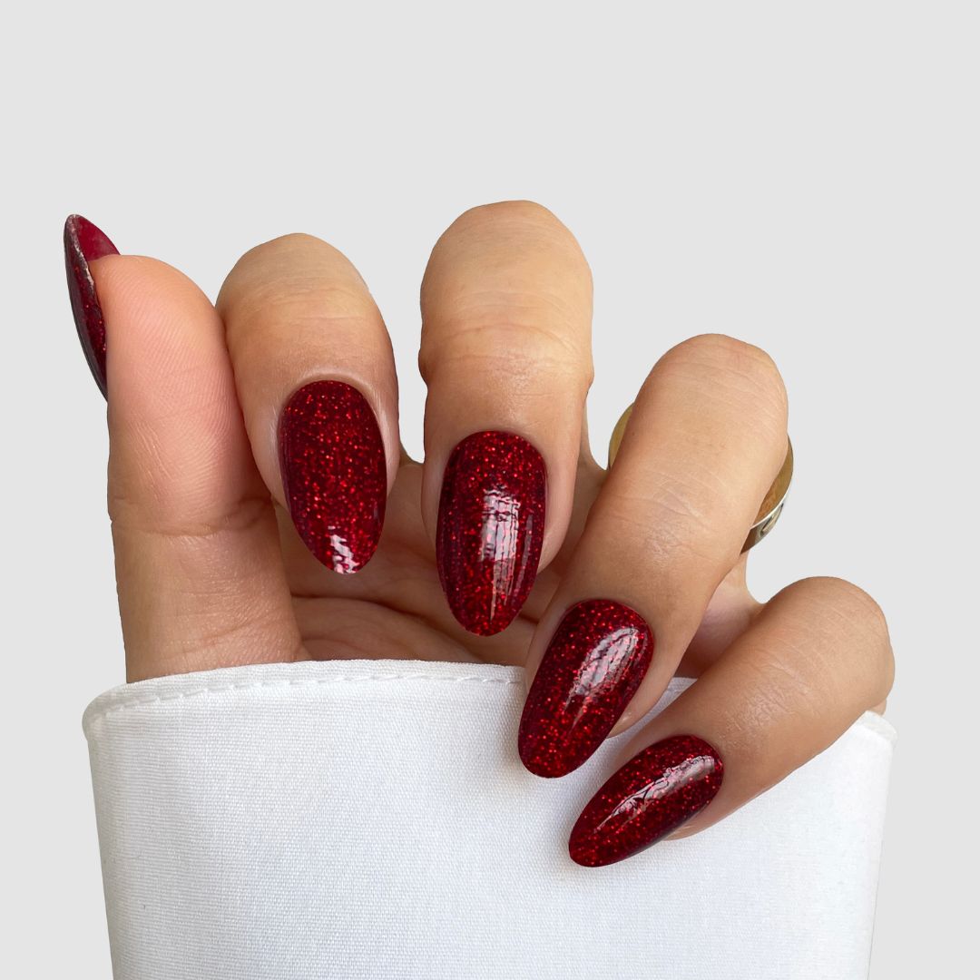 A hand with long, almond-shaped nails, featuring Sassy Saints’ Sparkling Ember Gel Wrap in deep red glitter, peeks from under a white sleeve.