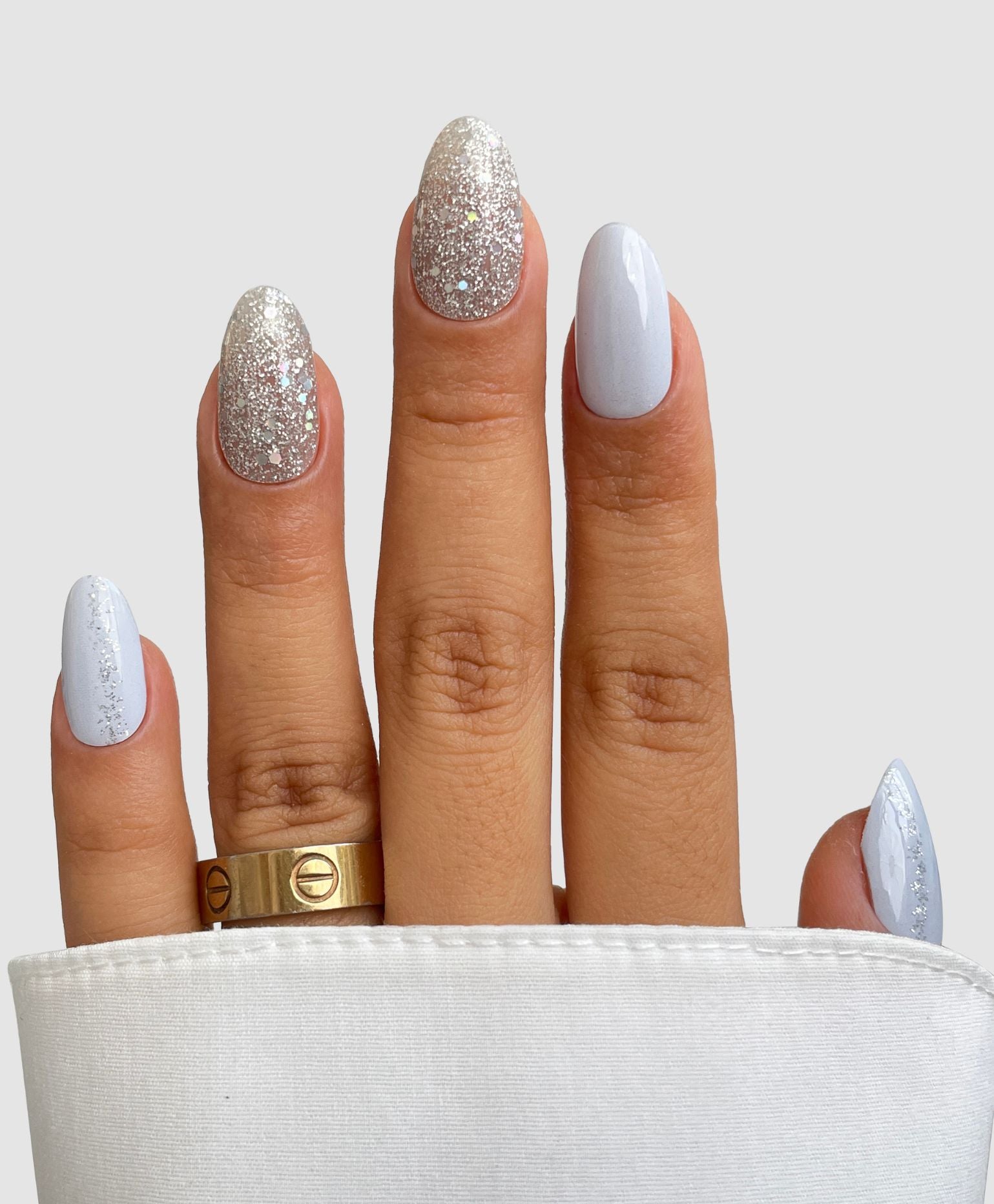 A hand with almond-shaped nails features Sassy Saints’ Hazy Frost Gel Wrap, alternating light blue and silver glitter. Two gold rings adorn the middle finger, and part of the hand is covered by a white sleeve.