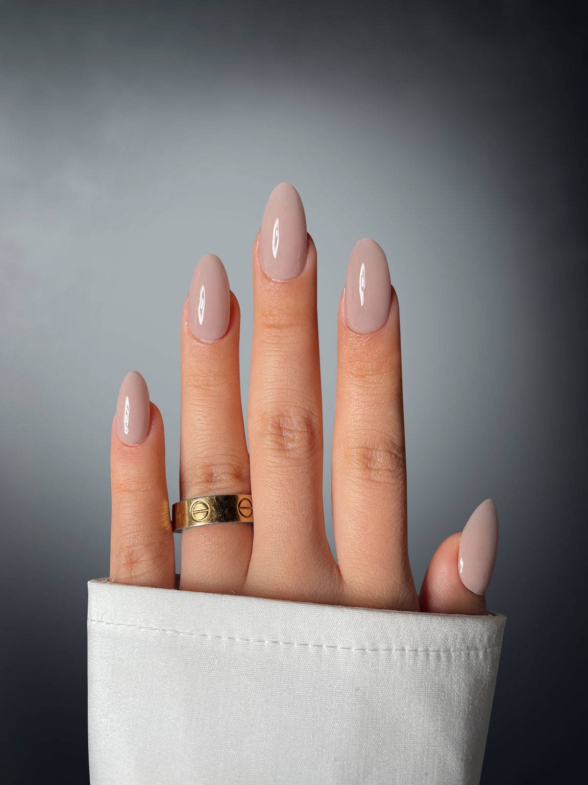 A hand with neatly manicured oval nails in Sassy Saints' Rosé Rebel Dipping Powder, wearing a gold ring on the middle finger and partially covered by a white cuff, is set against a gradient gray background.