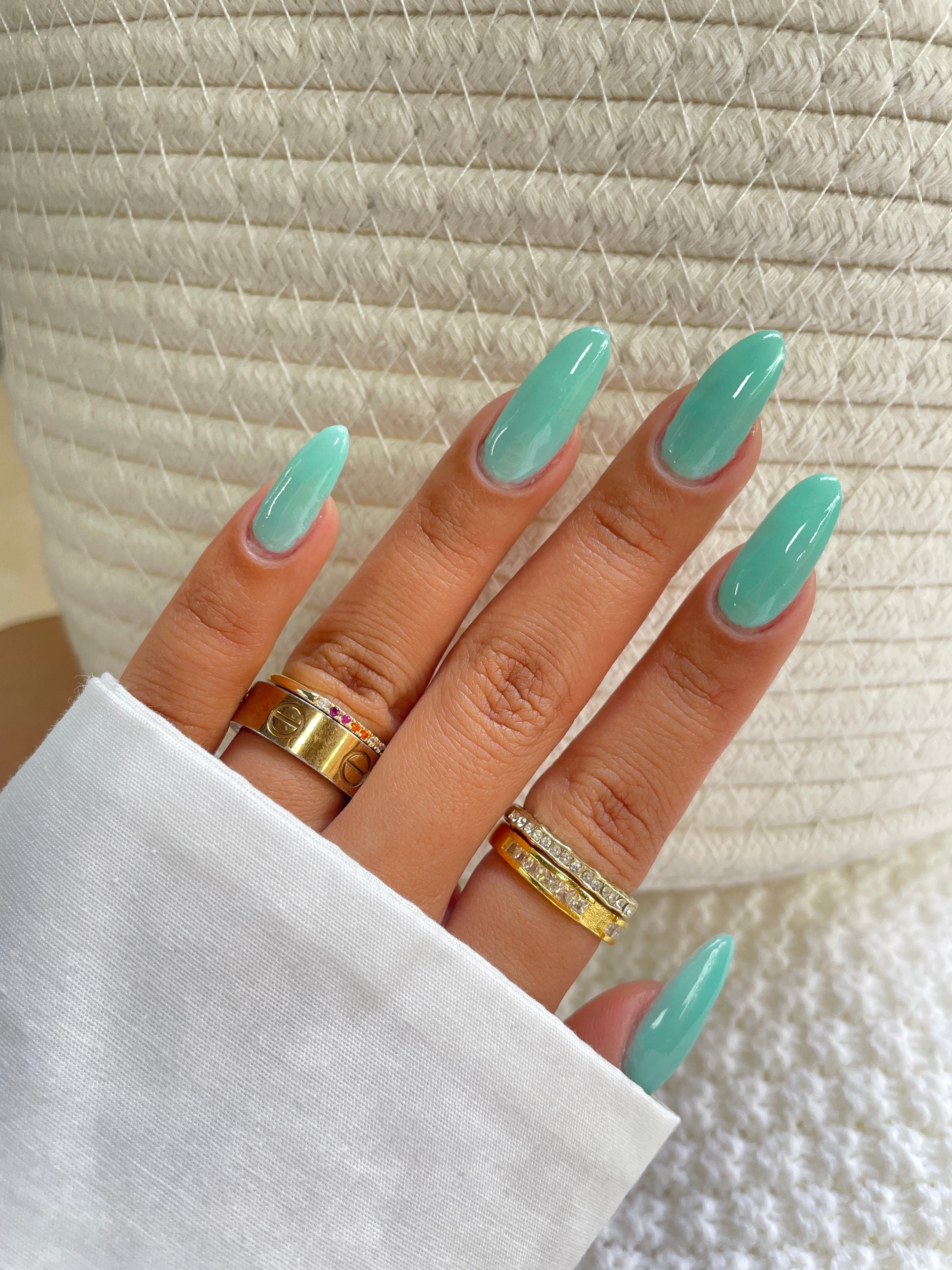 A hand with long, glossy teal nails—created using Sassy Saints’ Teal Tactician Dipping Powder—wears three gold rings and rests on a textured white surface, partially covered by a white sleeve.