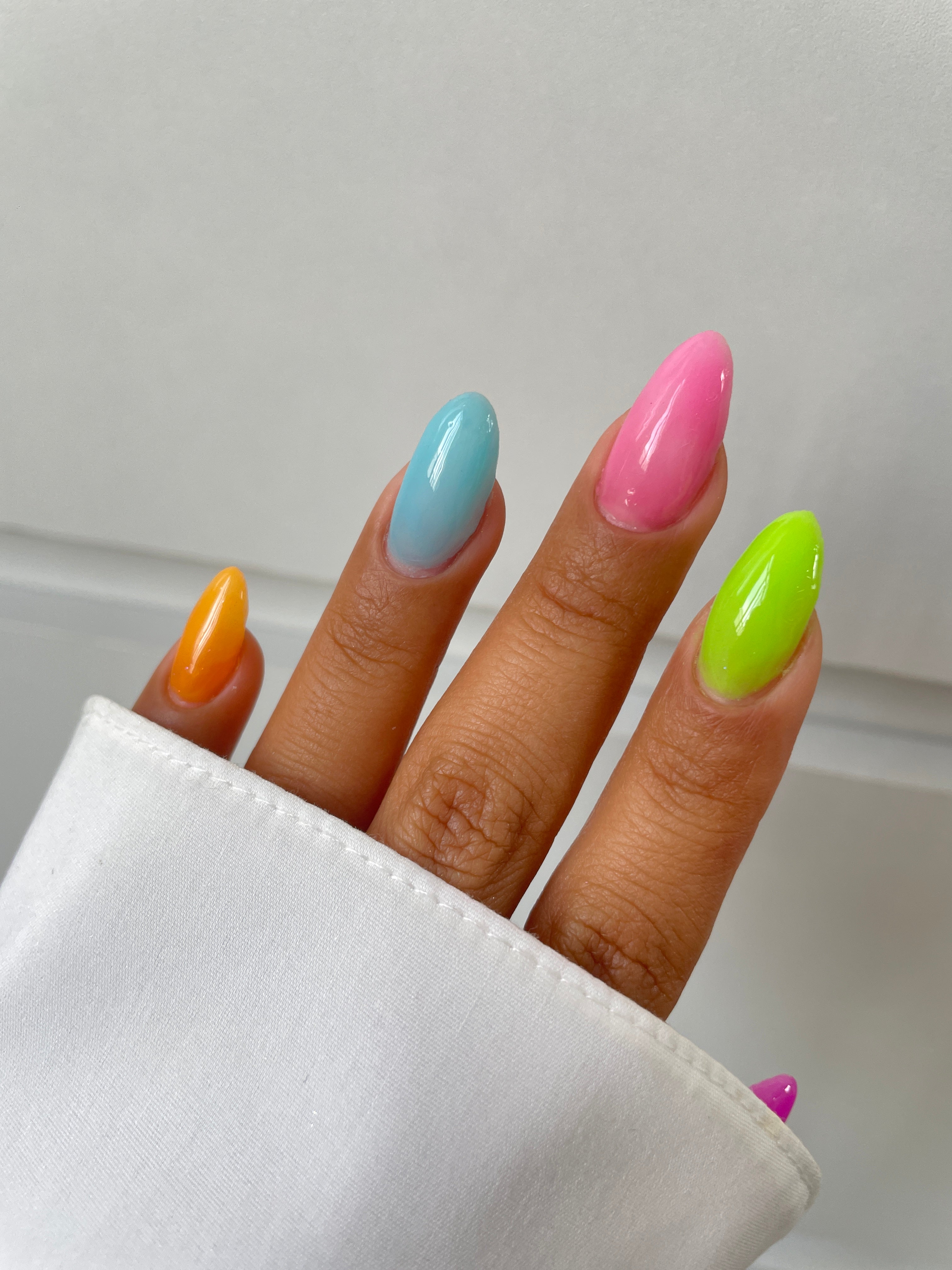 A hand with almond-shaped nails, painted in bright shades using Sassy Saints' Sunny Spritz Dipping Powder, is shown against a white background. The person wears a white sleeve, highlighting the vibrant colors and salon-quality finish.