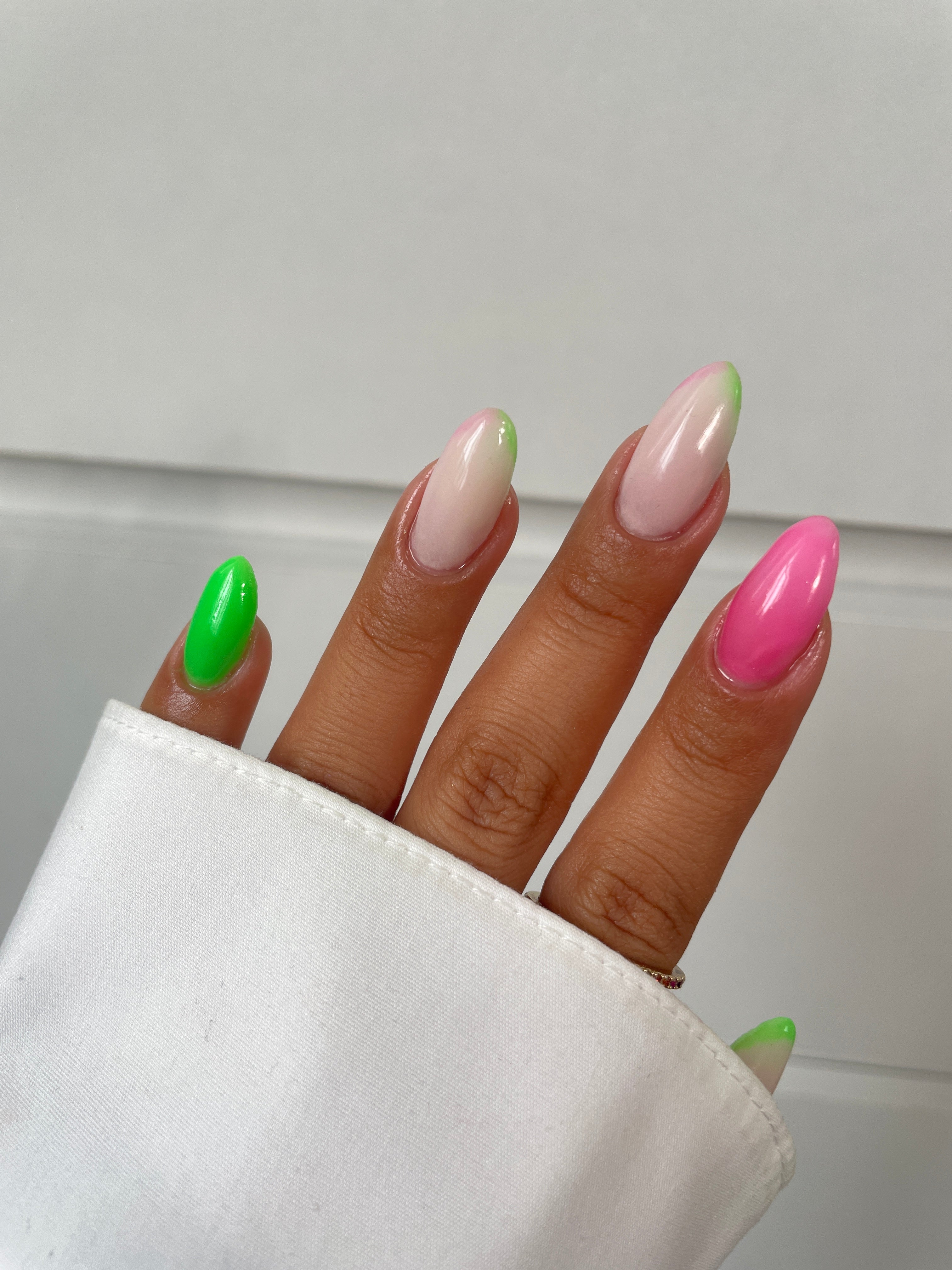 A hand with long almond-shaped nails showcases Sassy Saints' Palm Paradise Dipping Powder: one bright green, one bright pink, and others nude with neon tips—all partly hidden by a white sleeve. Brings The Salon Home.