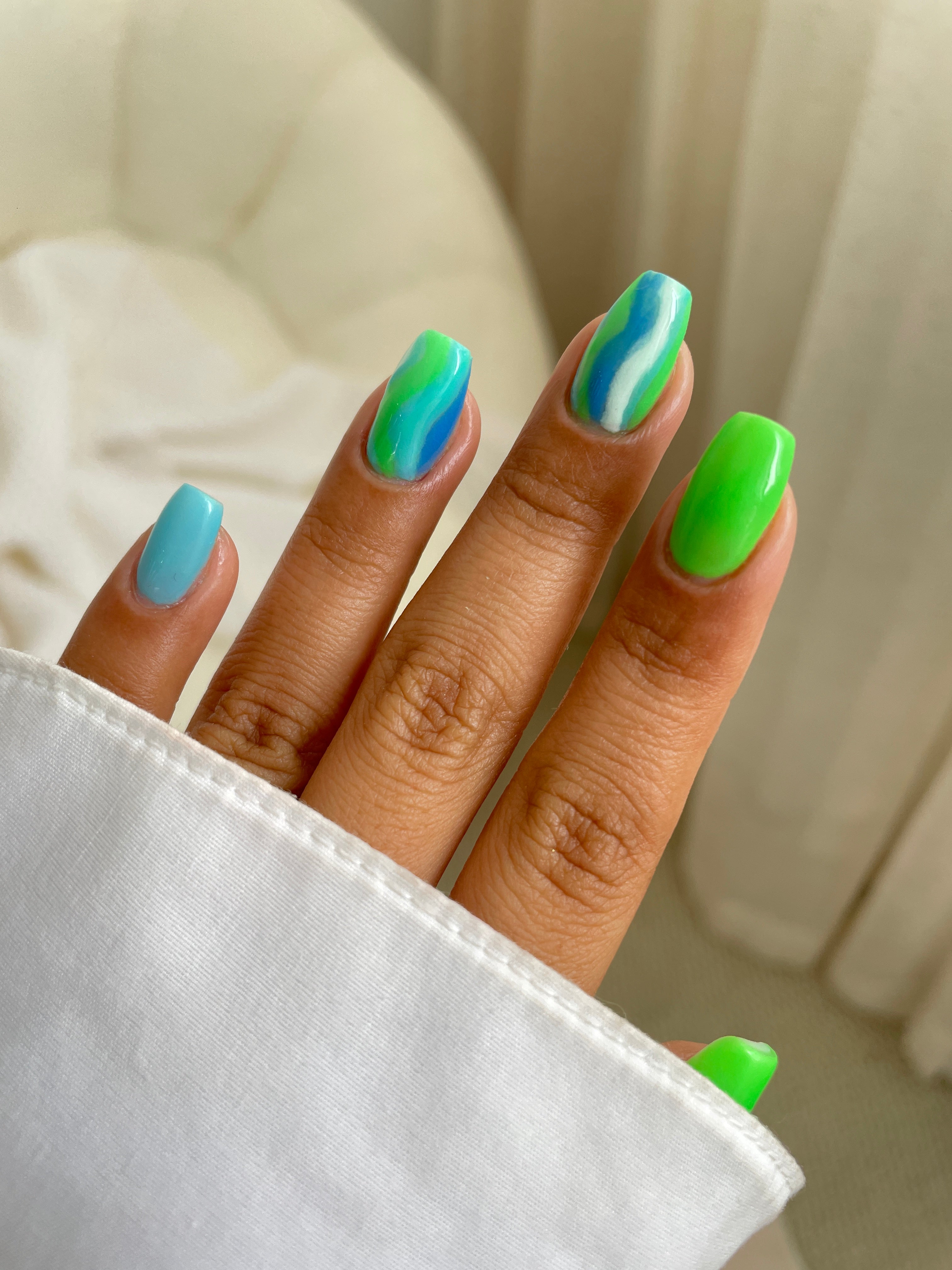 A hand with medium-length square nails features Sassy Saints' Blue Breeze Dipping Powder in bright blue and neon green, plus blue-green wavy art on the middle and ring fingers. A white sleeve and soft, neutral background complete the look.