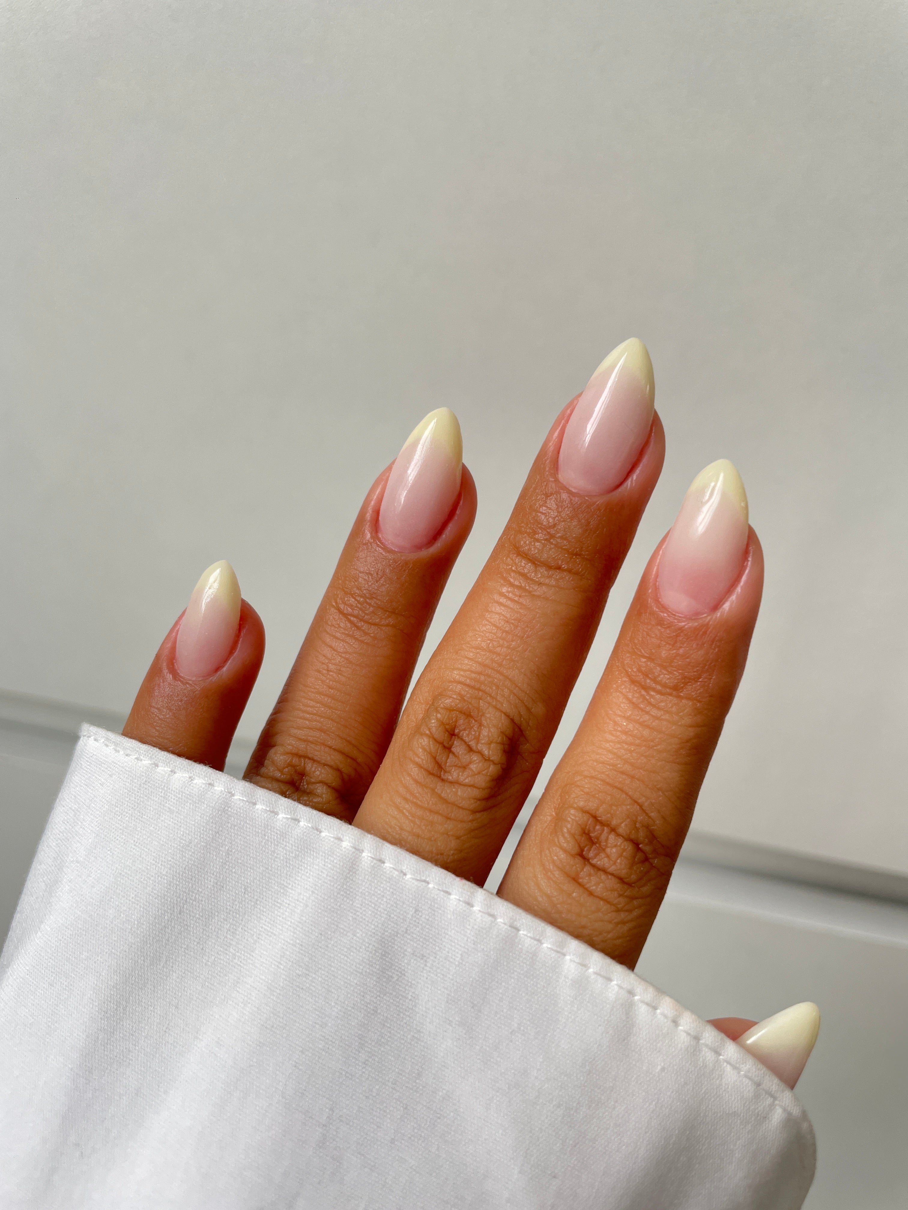 A hand with almond-shaped nails features the pale, natural ombre of Sassy Saints' Vanilla Veil Dipping Powder, blending pink bases into creamy white tips. The person wears a white long-sleeve shirt.