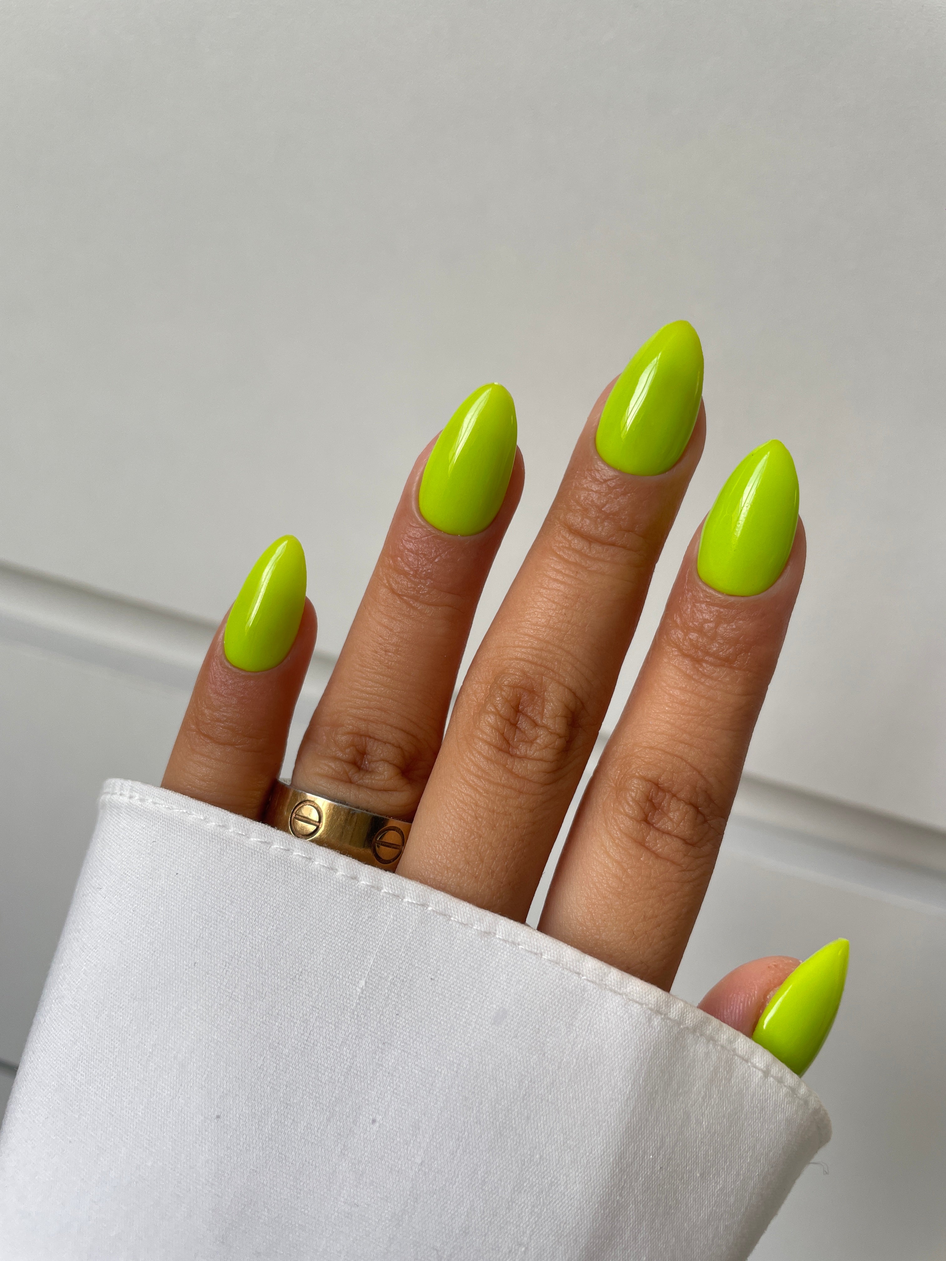 A hand with long, almond-shaped nails in Sassy Saints' Lime Luminary Dipping Powder, a neon green shade, rests against a white background. The look is accented by a gold ring and a partially visible white sleeve.