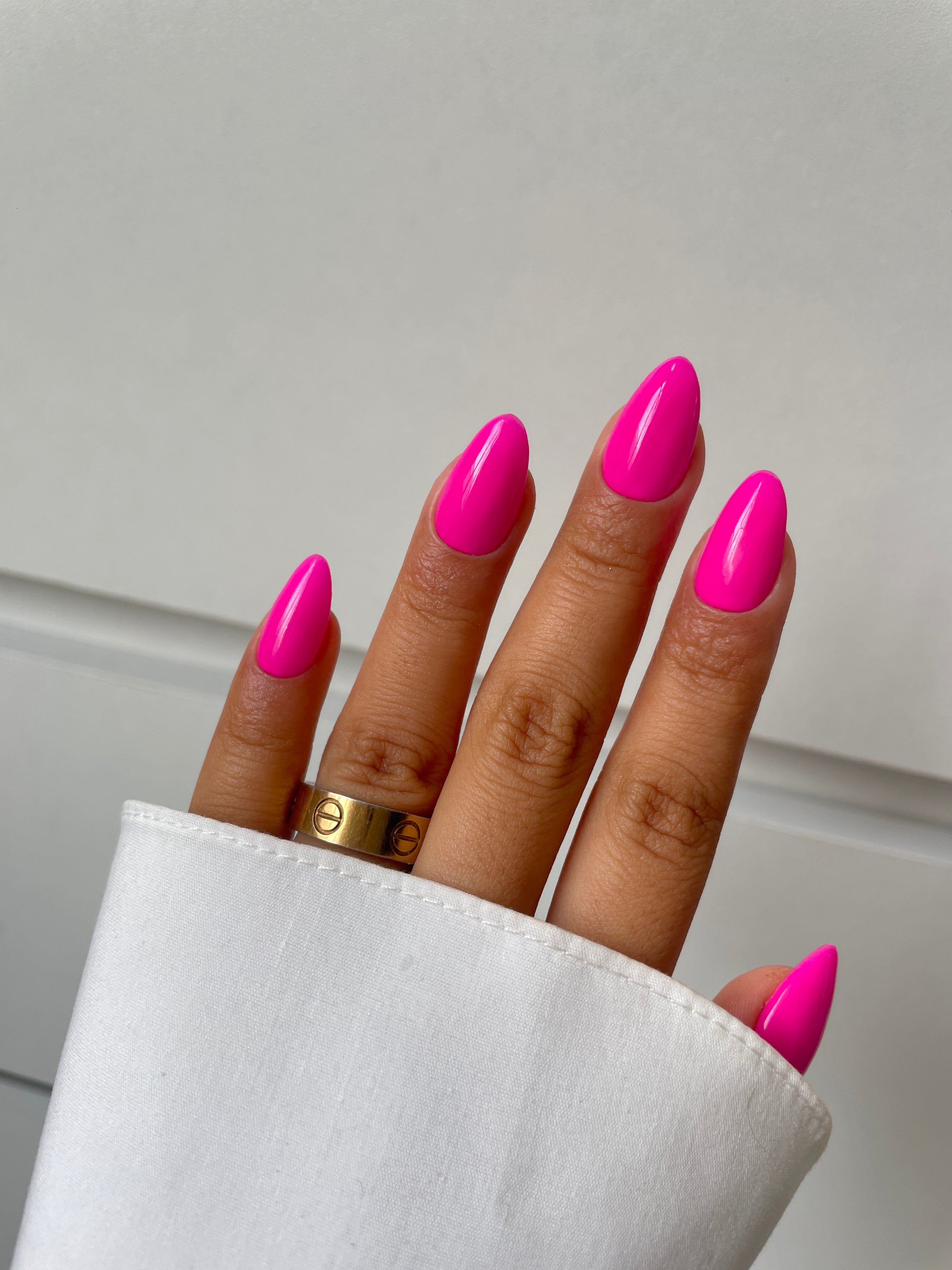 A hand with bright pink, almond-shaped nails showcases Sassy Saints' Daiquiri Dream Dipping Powder; a gold ring adorns the middle finger while a white sleeve partially covers the hand.