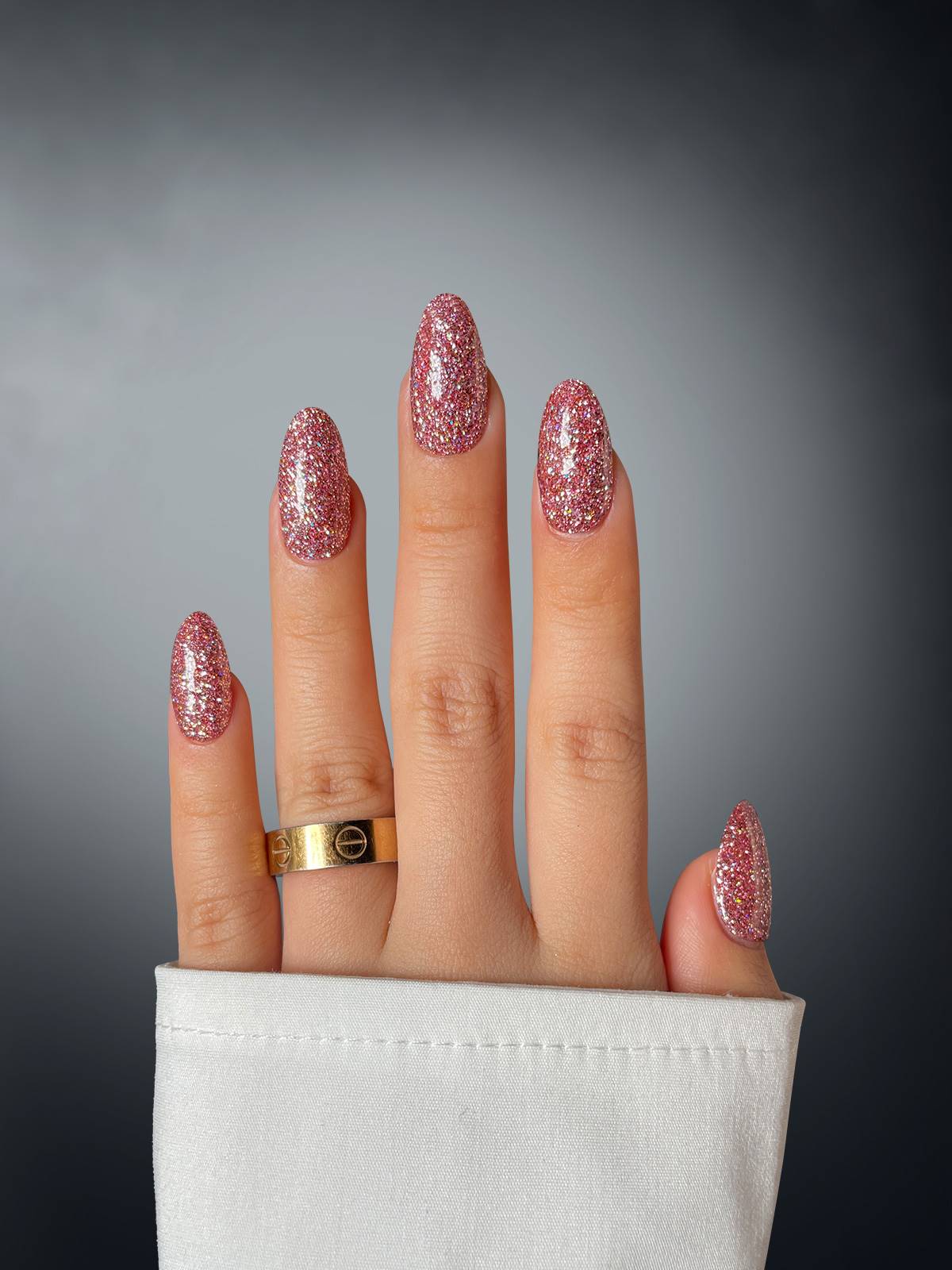 A hand with long, oval nails painted in Sassy Saints - Brings The Salon Home Sparkly Spirited Dipping Powder. A gold ring adorns the ring finger, paired with a white sleeve. Background features a dark to light gray gradient.