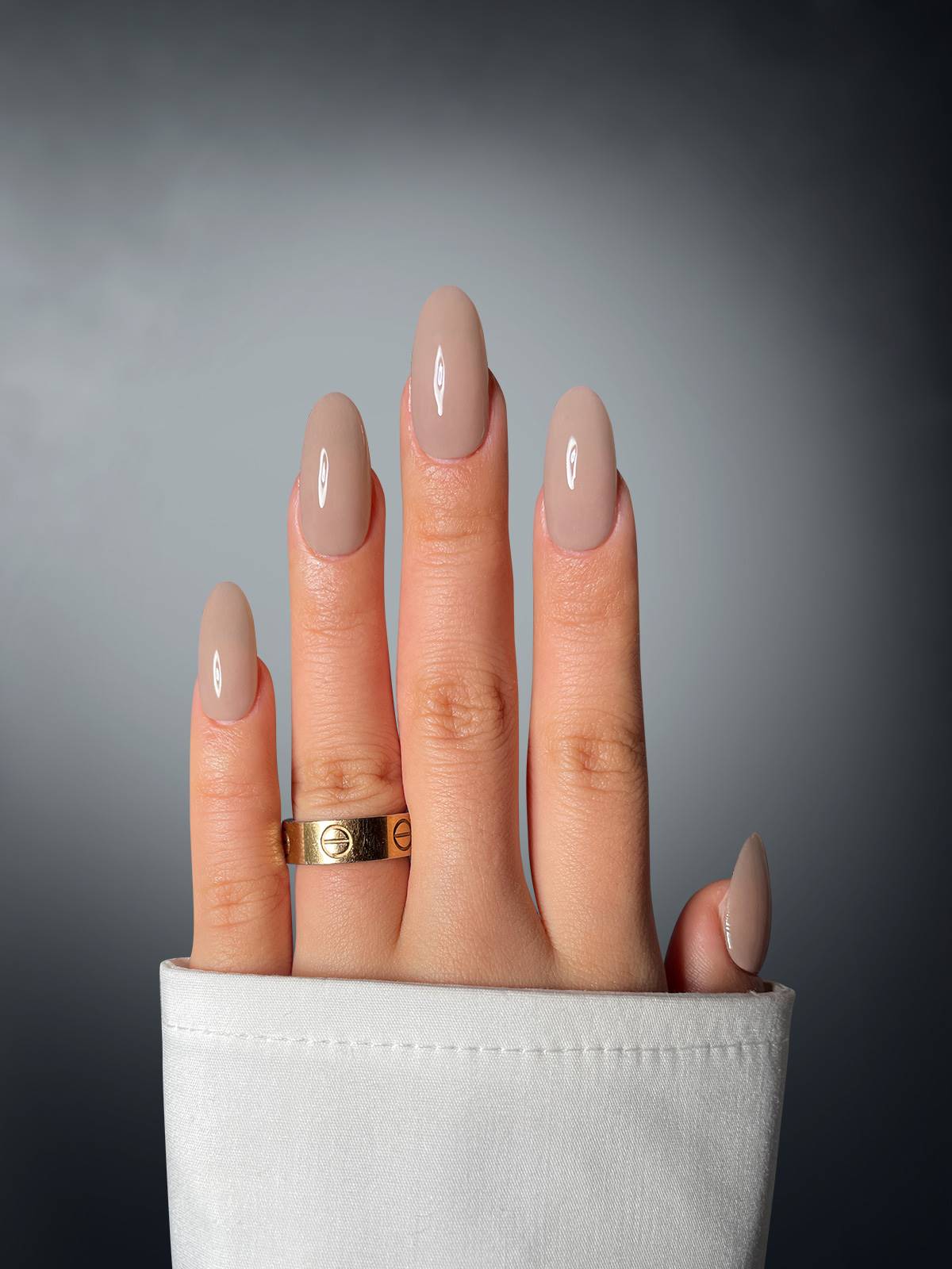 A hand with neatly manicured, nude nails using Sassy Saints' Radiant Rebel Dipping Powder wears a gold ring and rests against a gray background, partially covered by a white sleeve.