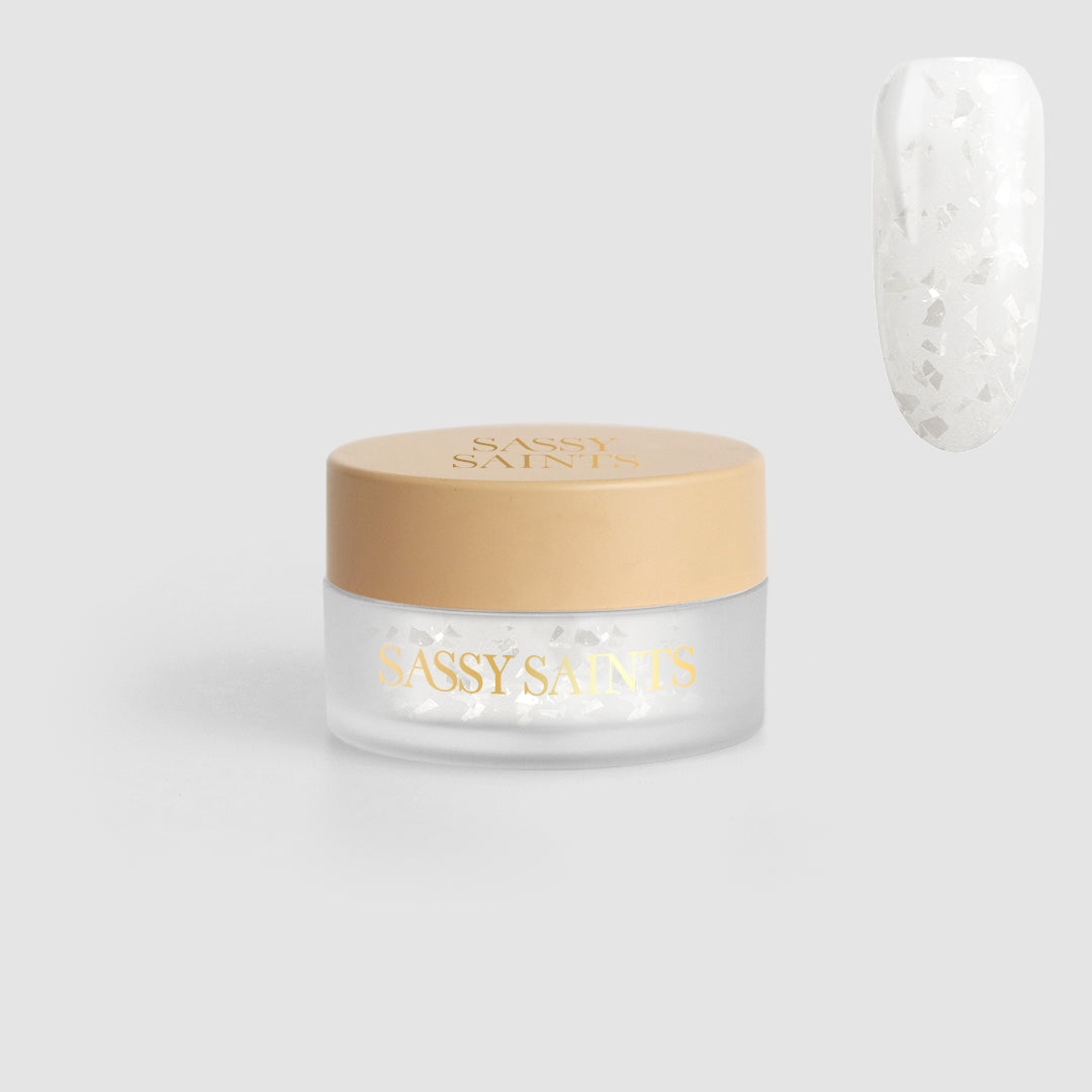 A small round jar labeled Winter's Whisper Dipping Powder from Sassy Saints - Brings The Salon Home, with a beige lid, contains white nail flakes. A sample nail shows the flakes on a clear base. The background is light gray.