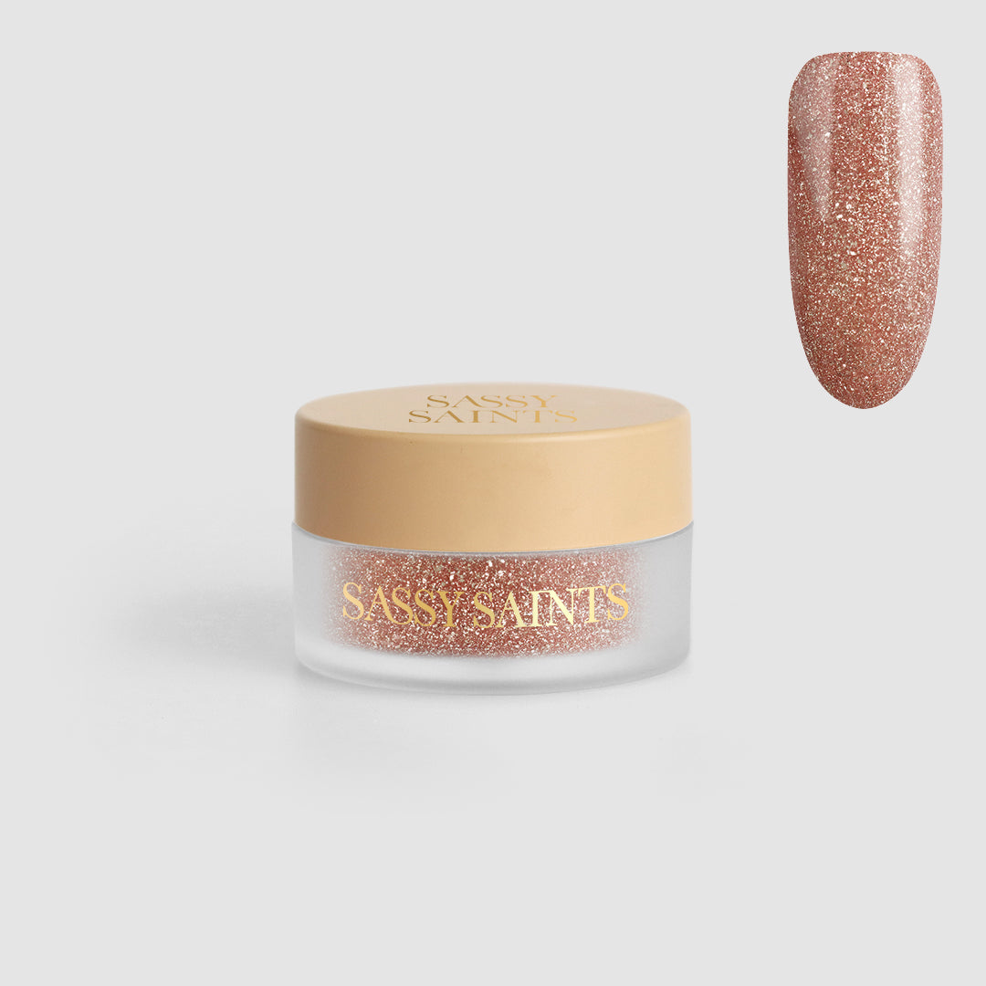A small round jar labeled “Toasted Tinsel Dipping Powder” from Sassy Saints – Brings The Salon Home holds rose gold glitter powder. Above and right, a nail swatch displays its sparkling rose gold finish on a light gray background.