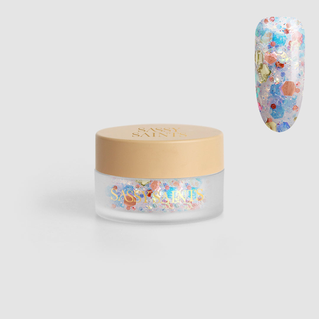 A small clear jar with a beige lid labeled Sassy Saints - Brings The Salon Home, containing Fairy Fizz Dipping Powder. A sample nail displays the powder in pastel colors and gold accents for a colorful, salon-quality look.