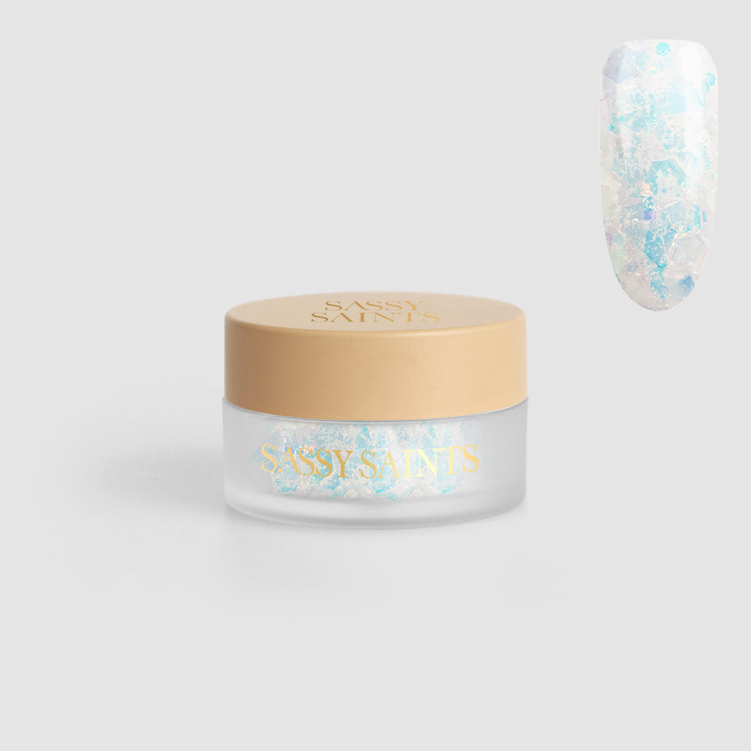 A small clear jar with a beige lid labeled Sassy Saints - Brings The Salon Home, containing Cotton Candy Confetti Dipping Powder. A sample shows the powder applied, creating a shimmery, opalescent effect on a nail.