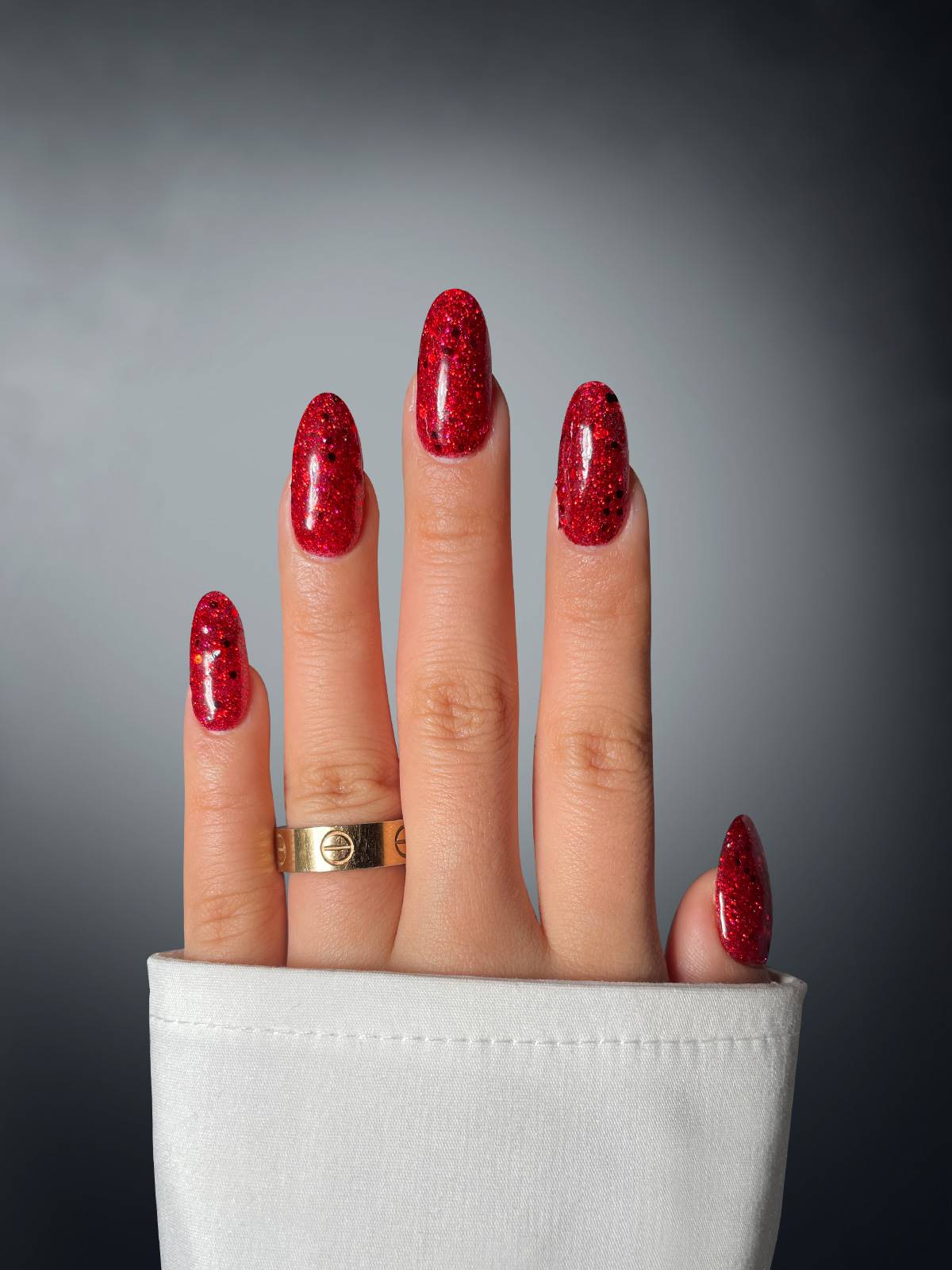 A hand with almond-shaped nails features Sassy Saints' Sultry Siren Dipping Powder, a red glitter polish, against a dark gradient background. A gold ring and white cuffed sleeve add to the look. Brings The Salon Home.