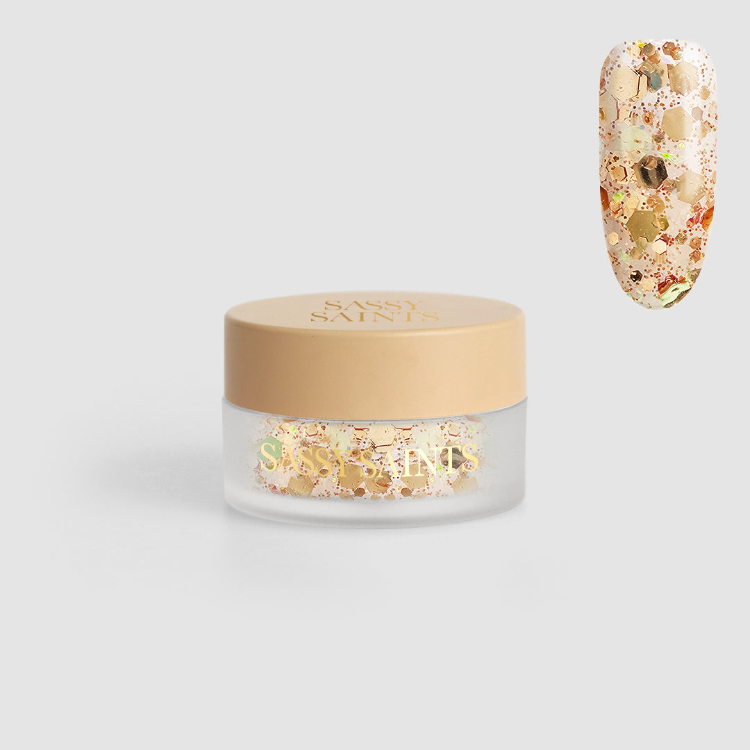 A small clear jar with a beige lid labeled Sassy Saints - Brings The Salon Home contains Honey Glaze Dipping Powder. Above and to the right is a close-up of a fingernail decorated with chunky gold glitter from the powder.
