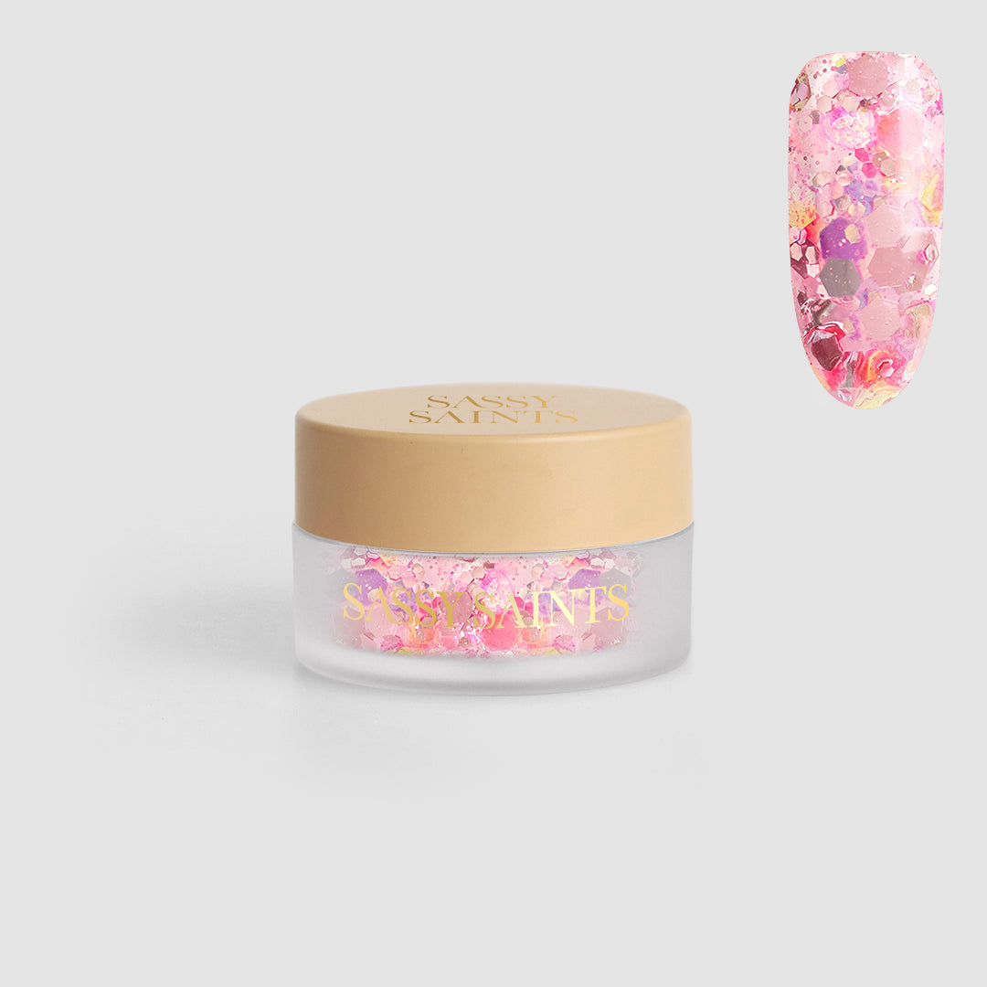 A small clear jar with a beige lid labeled "Sassy Saints - Brings The Salon Home" holds Spring Rose Dipping Powder; a sample nail displays this pink and iridescent powder in a colorful, sparkling pattern.