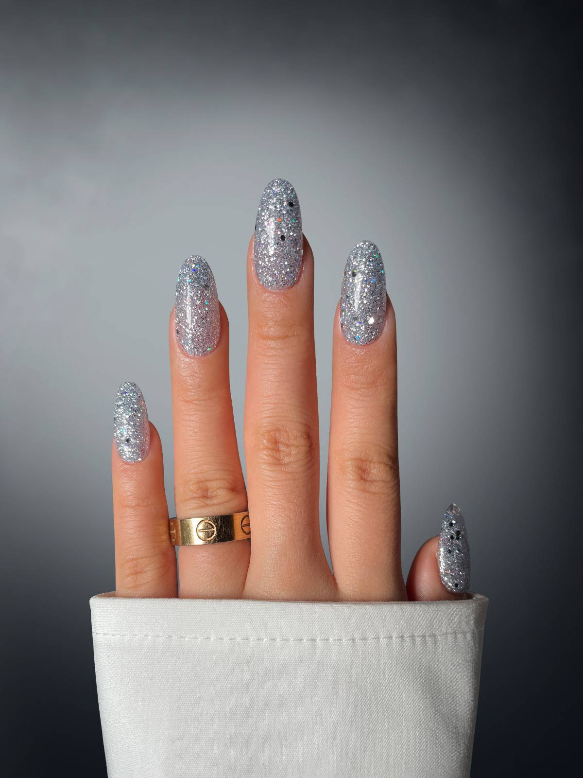 A hand with long, oval nails coated in Sassy Saints’ Foggy Flickerer Dipping Powder, adorned with a gold ring and white sleeve, stands out against a dark gradient background.