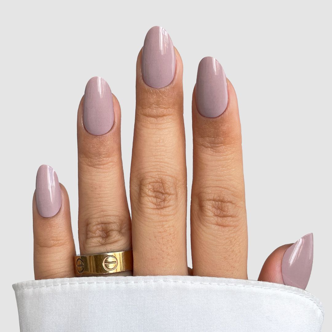 A hand features long, almond-shaped nails with Sassy Saints’ Neutral Flow Gel Wrap in a glossy pale mauve. A gold ring adorns the middle finger, while a white fabric partially covers the hand. Brings The Salon Home.